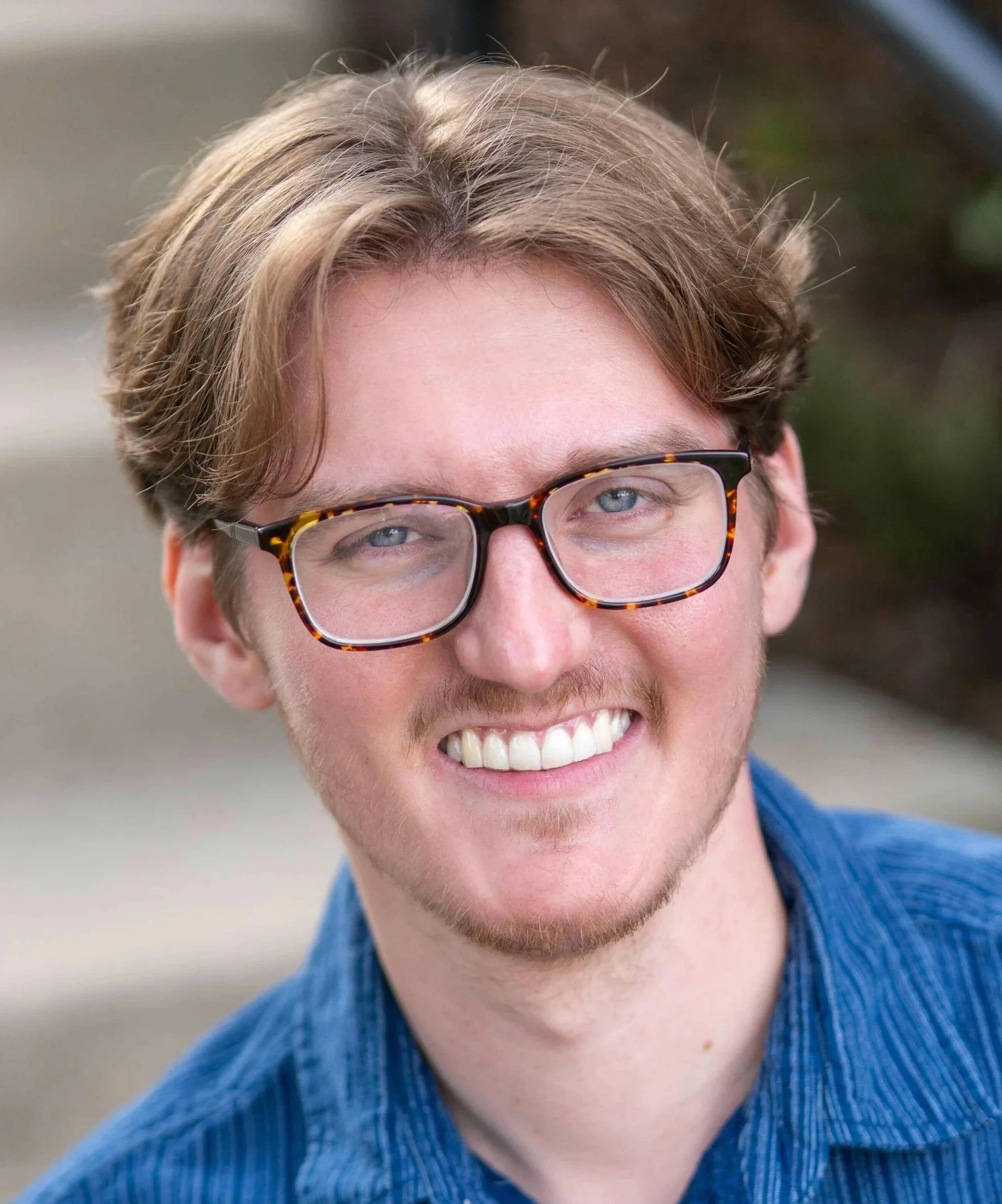 Close-up of a smiling young man with light brown hair, wearing tortoiseshell glasses and a blue sweater, outdoors in daytime.