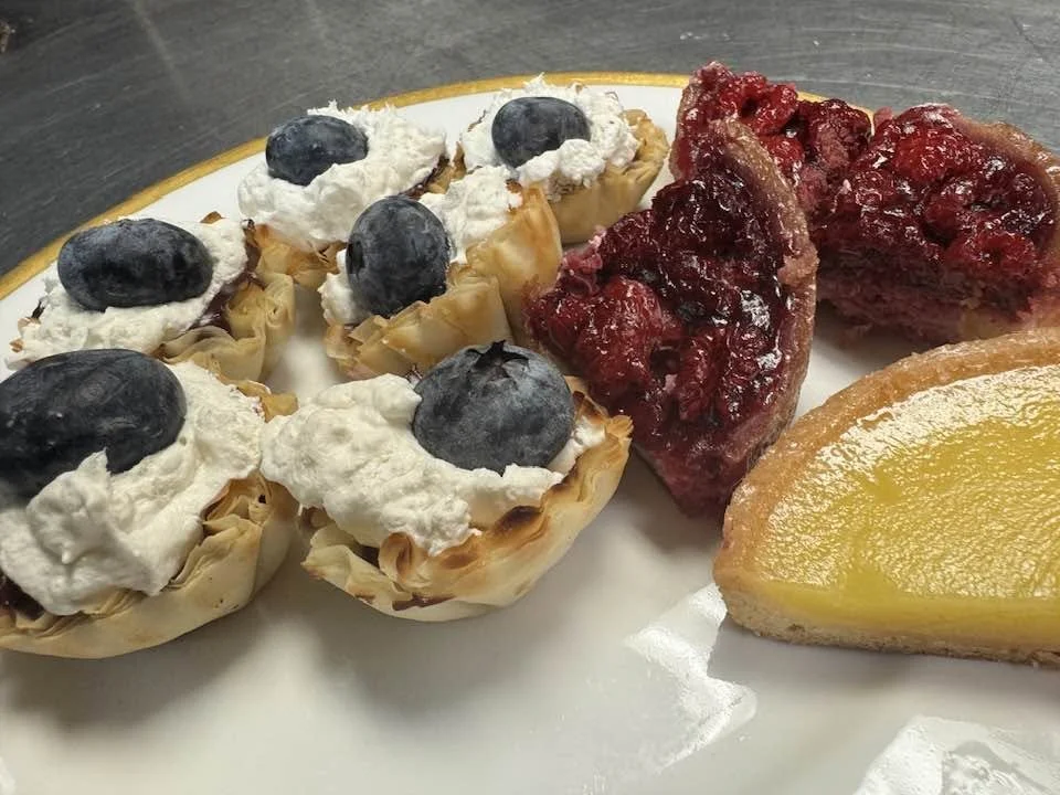Three mini taco shells filled with whipped cream and topped with blueberries, a slice of cherry dessert, and a slice of lemon tart on a white plate.