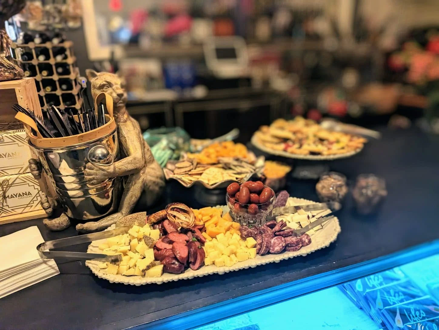 Charcuterie board with cheese, meats, olives, and crackers, with a decorative, metallic frog sculpture holding utensils on the side, set on a counter with a dark tablecloth.