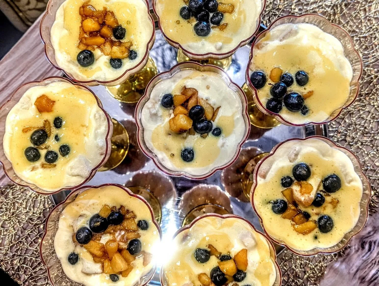 Multiple glasses filled with a creamy dessert topped with blackberries and diced peaches, arranged on a metallic tray.