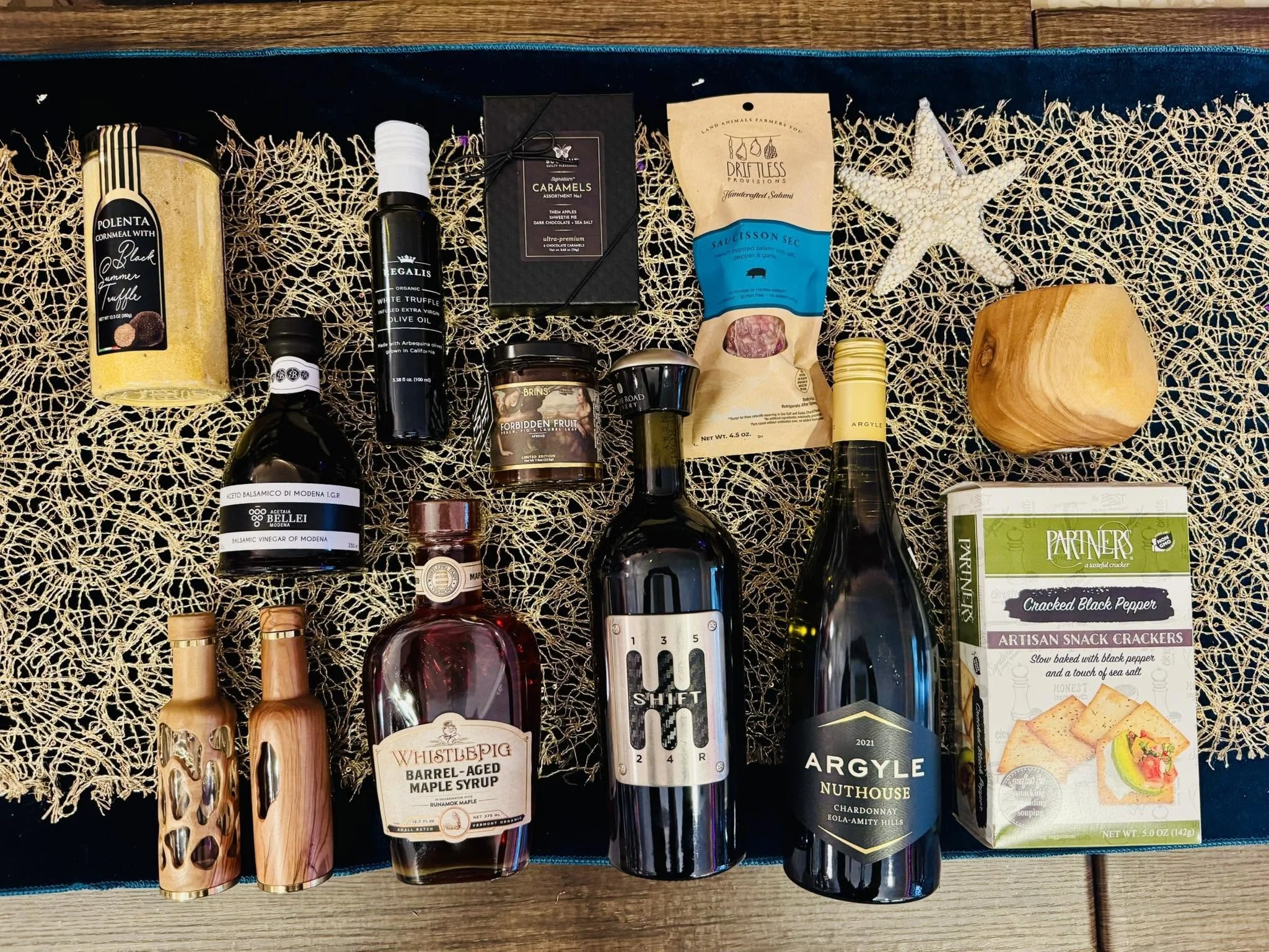 Arrangement of gourmet food items on a woven mat, including canned and bottled products, crackers, and decorative starfish.