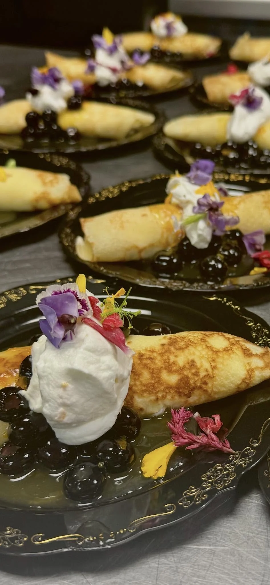 Crepes drizzled with chocolate syrup, topped with whipped cream, edible flowers, and fresh berries on ornate black plates.