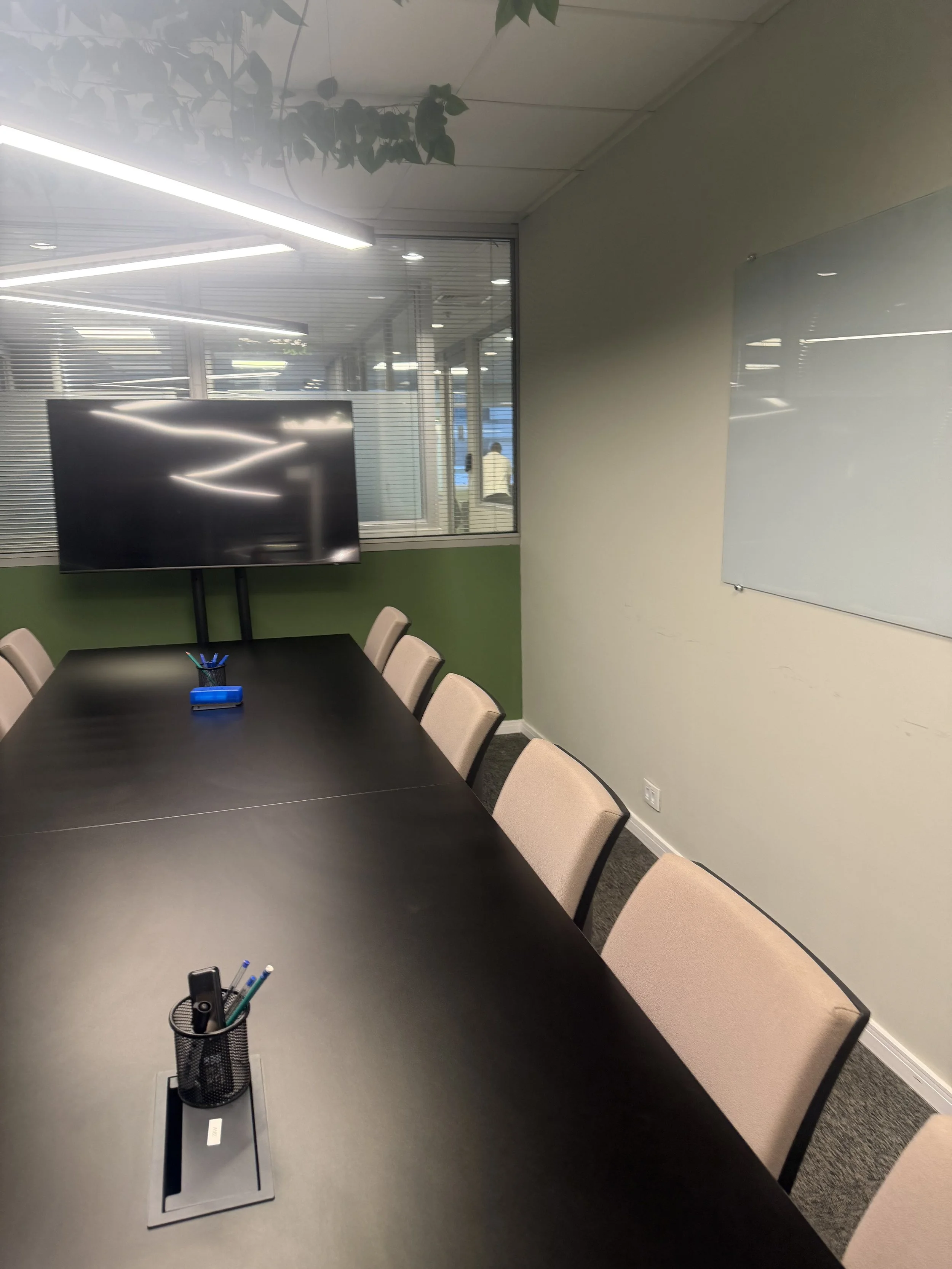 Sala de reunião com mesa preta, cadeiras bege, uma tela de TV na parede, uma janela com cortinas horizontais, uma pessoa sentada ao fundo, objetos de escritório na mesa, parede verde na parte inferior e branca acima, quadro branco na parede à direita.