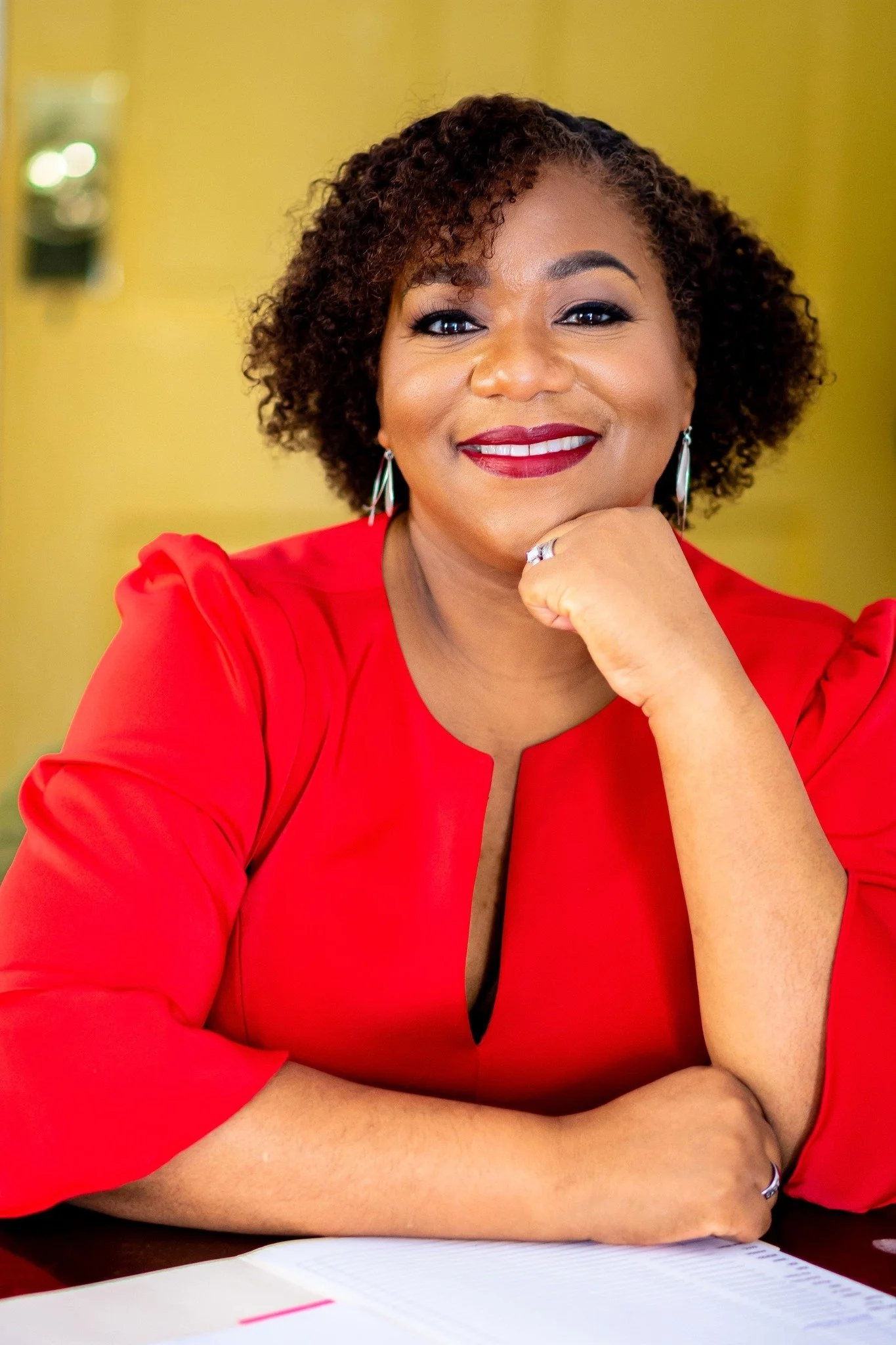 A woman with curly hair, wearing a red top and earrings, smiling and resting her chin on her hand, sitting at a desk with an open notebook.