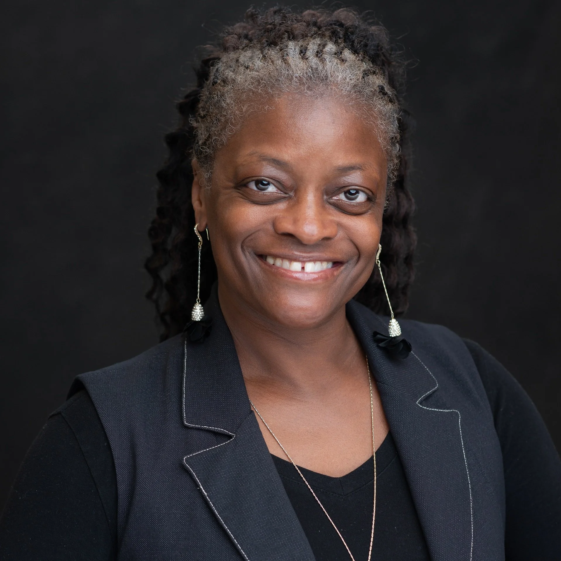 Close-up of a smiling Black woman with short, curly hair with gray streaks, wearing earrings and a black blazer with white piping, against a black background.