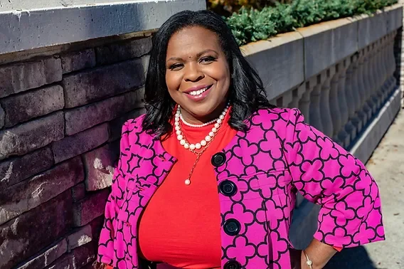 A woman standing outdoors with a brick wall and greenery in the background. She is smiling and wearing a pink patterned blazer, an orange top, and a pearl necklace.