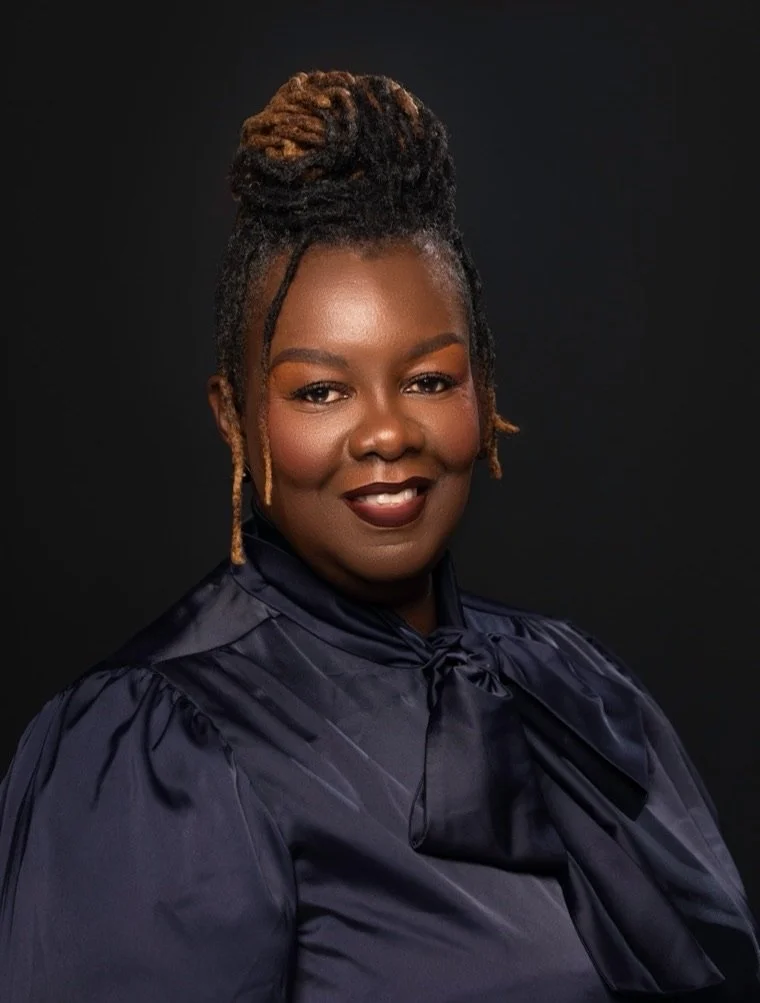 Portrait of a Black woman with styled dreadlocks in an updo, wearing a dark satin blouse with a bow at the collar, smiling against a dark background.