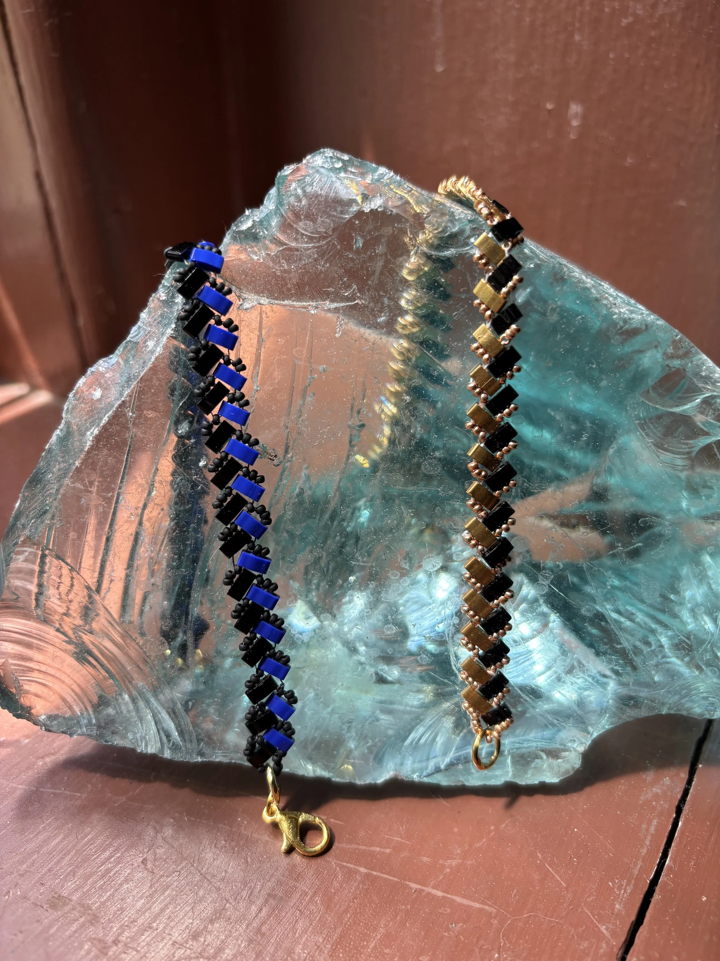 Two bracelets, one black and blue, the other black and gold, are draped over a clear, textured, and slightly curved glass object with some iridescent reflections. The background consists of a reddish-brown surface and a reddish-brown wall.