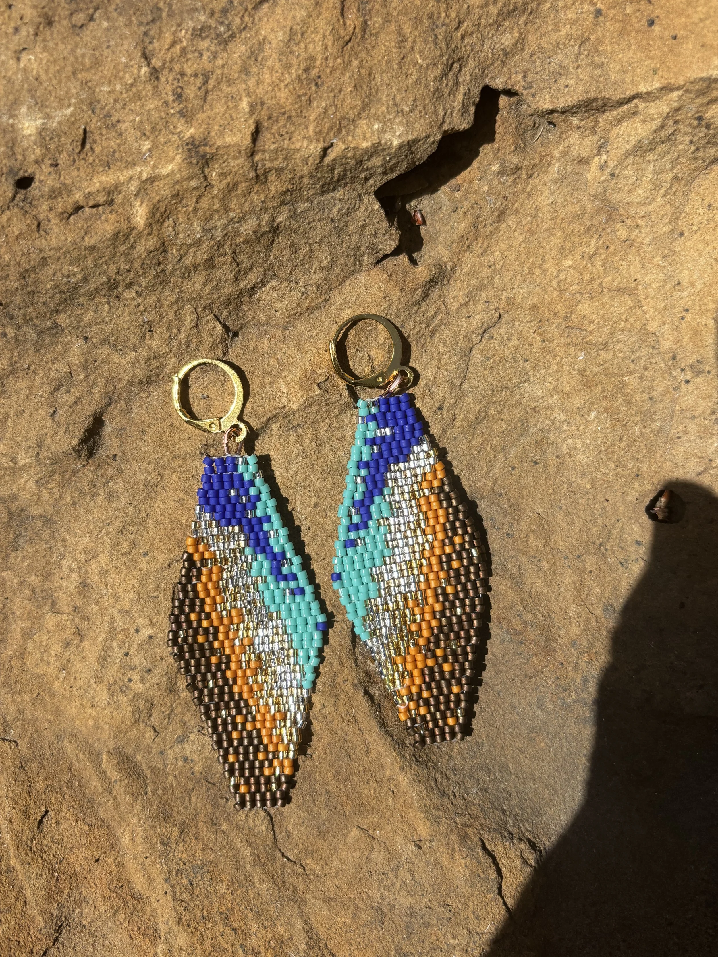 Colorful beaded earrings with a teardrop shape, hanging on a rough, brown rock surface.