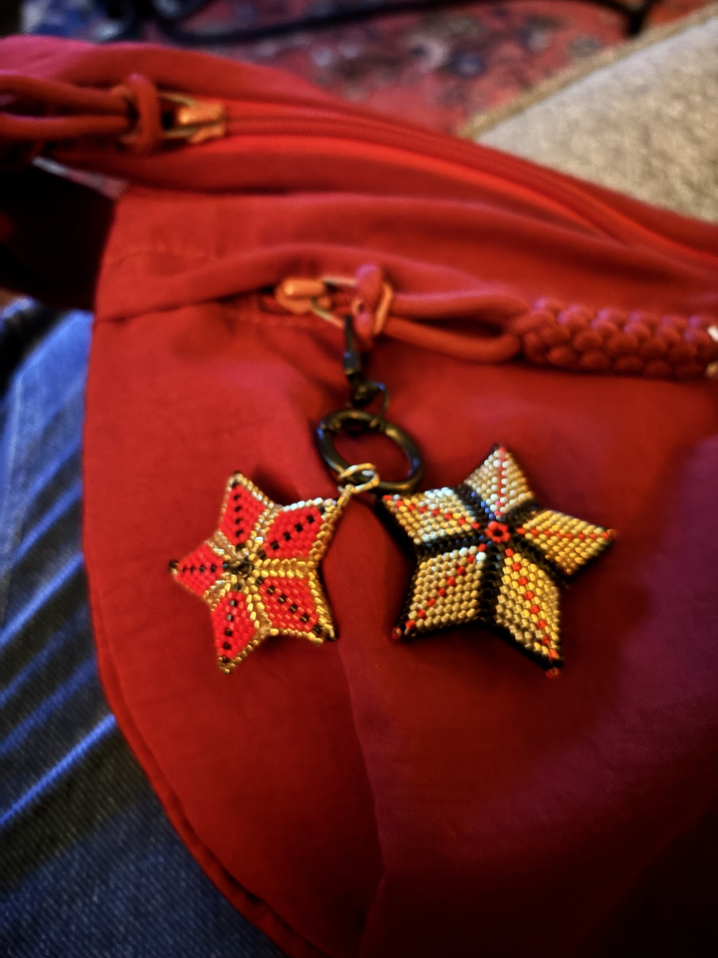 Close-up of two beaded star-shaped pendants, one red and black, the other yellow and black, attached to a keychain with a black metal ring. The background appears to be red fabric and part of a textured surface.