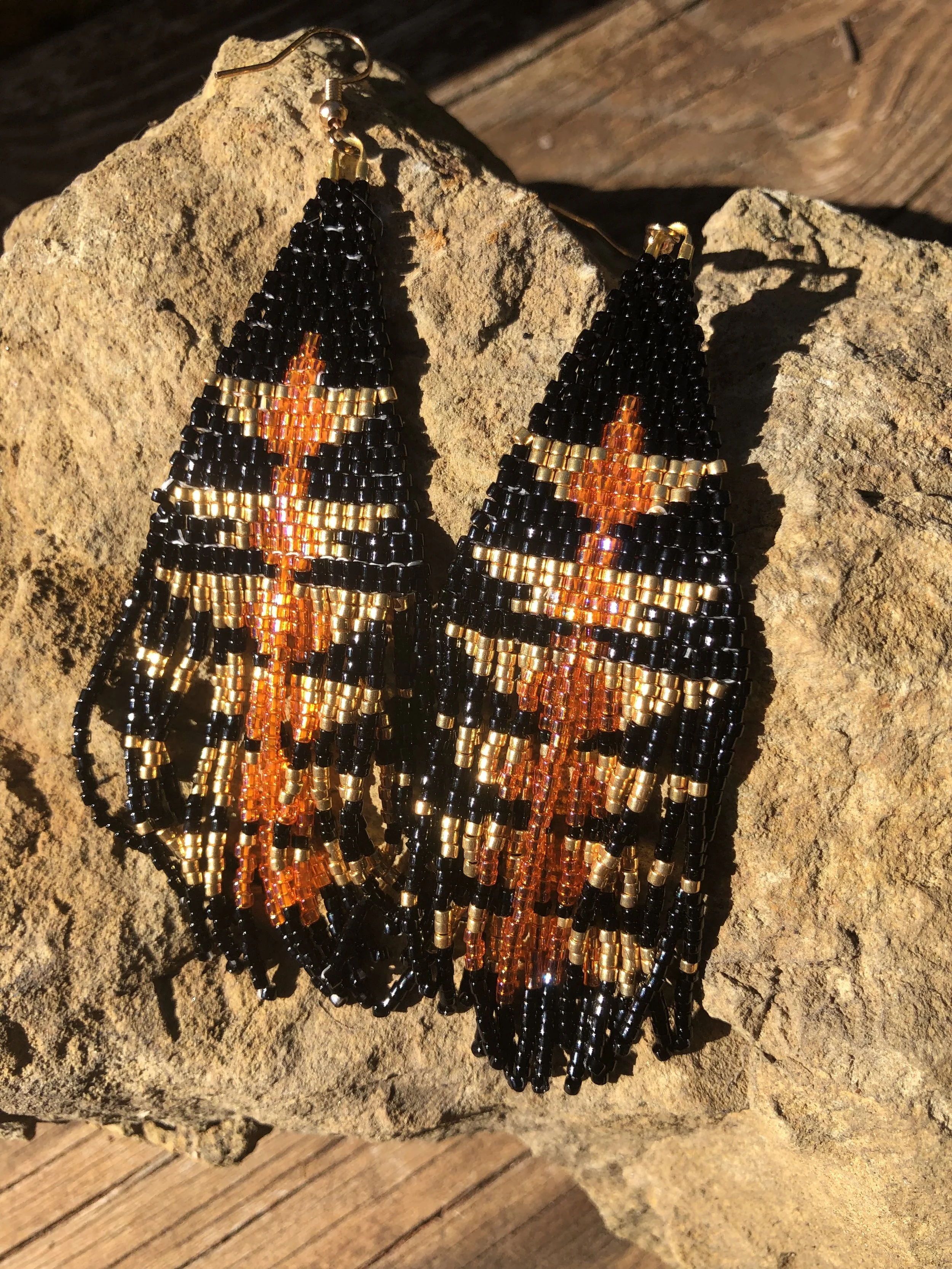 Beaded earrings with a diamond-shaped pattern in black, gold, and orange colors, resting on a rock.