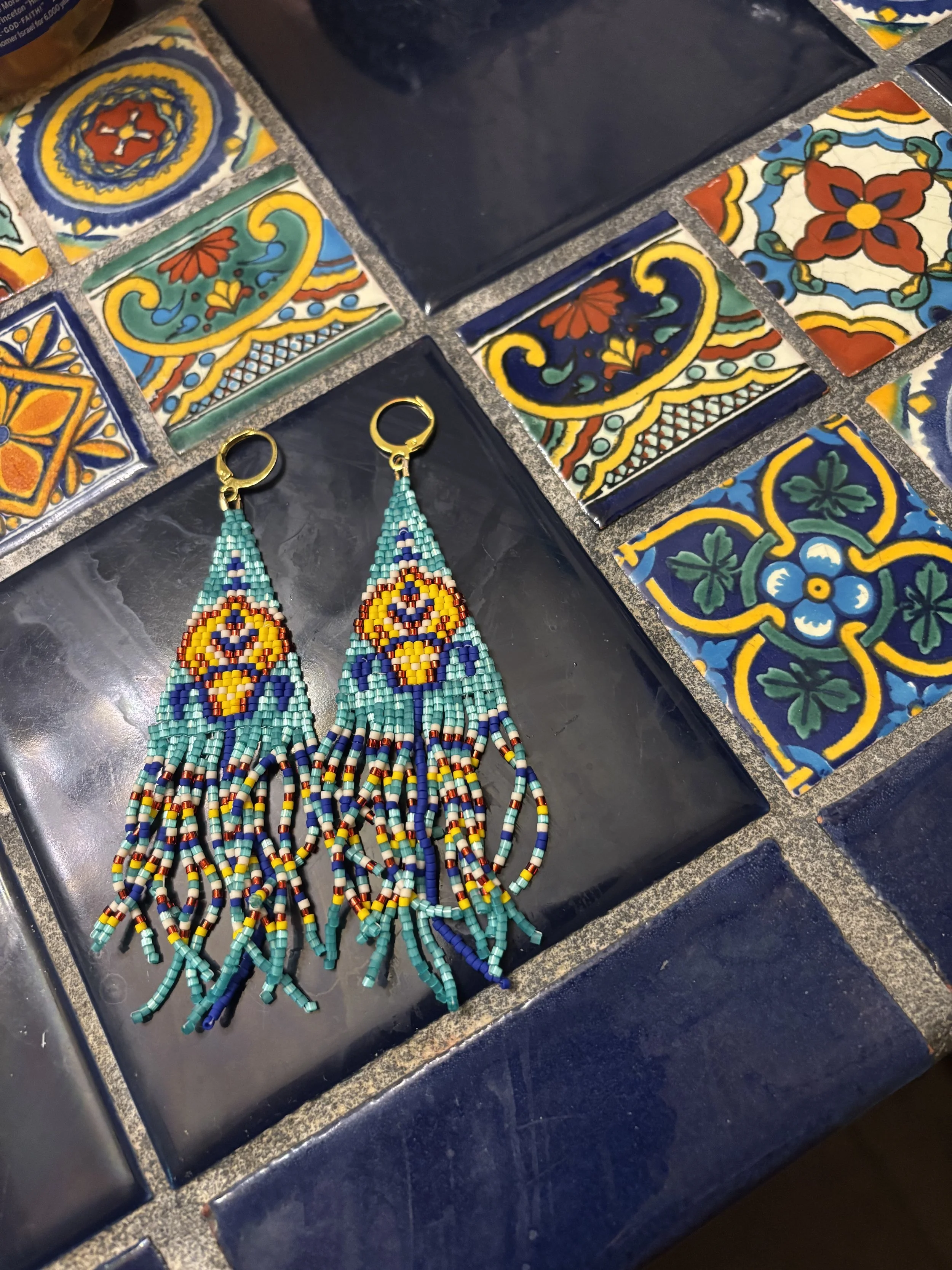 Colorful beaded earrings with intricate patterns laid out on a black surface surrounded by decorative ceramic tiles with vibrant patterns.
