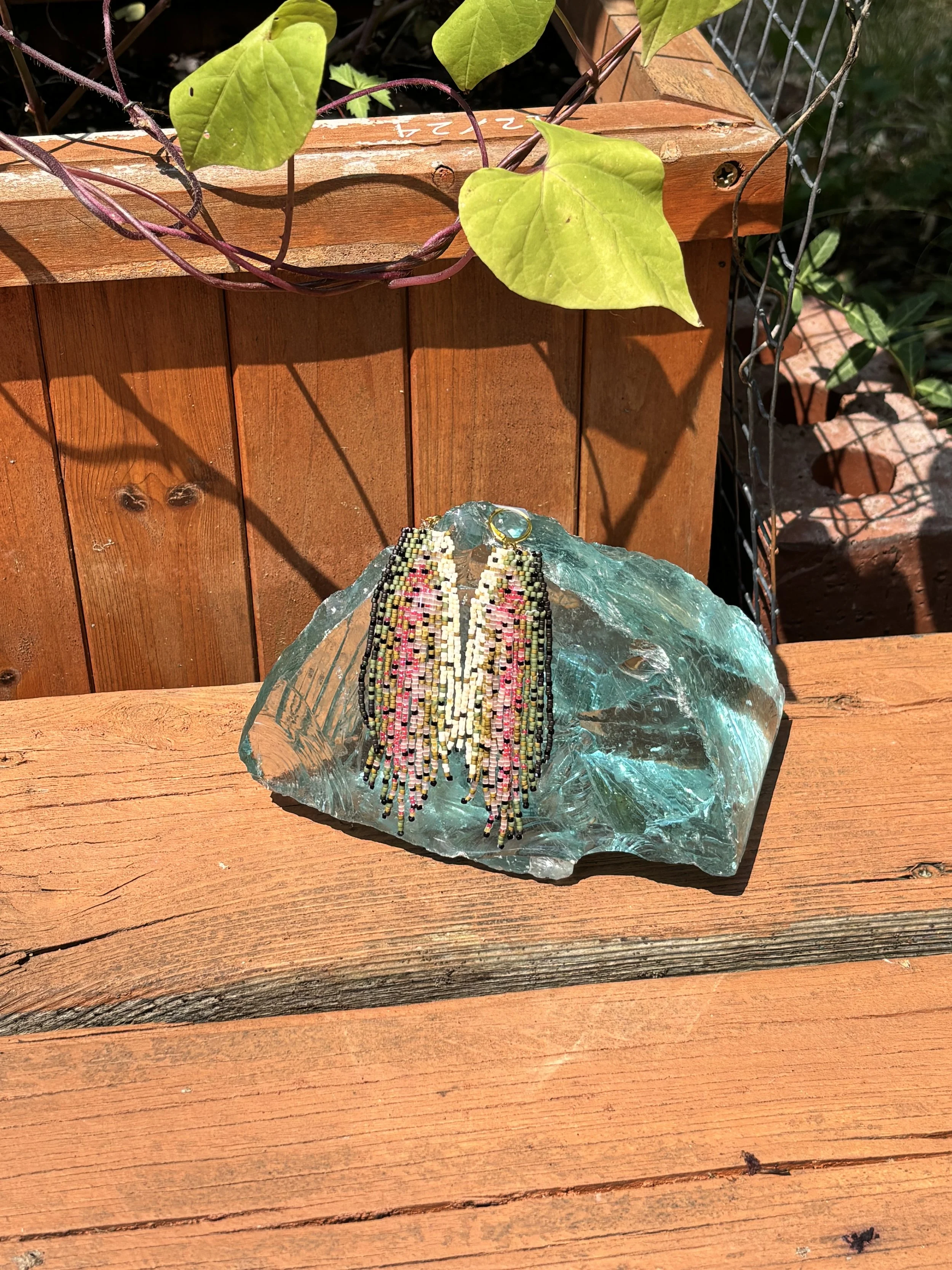 A small, beaded necklace with pink, yellow, white, and black beads, resting on a large, textured, light blue-green rock. The rock is on a wooden surface outside, with a wooden planter box, some green leaves, and a chain-link fence in the background.