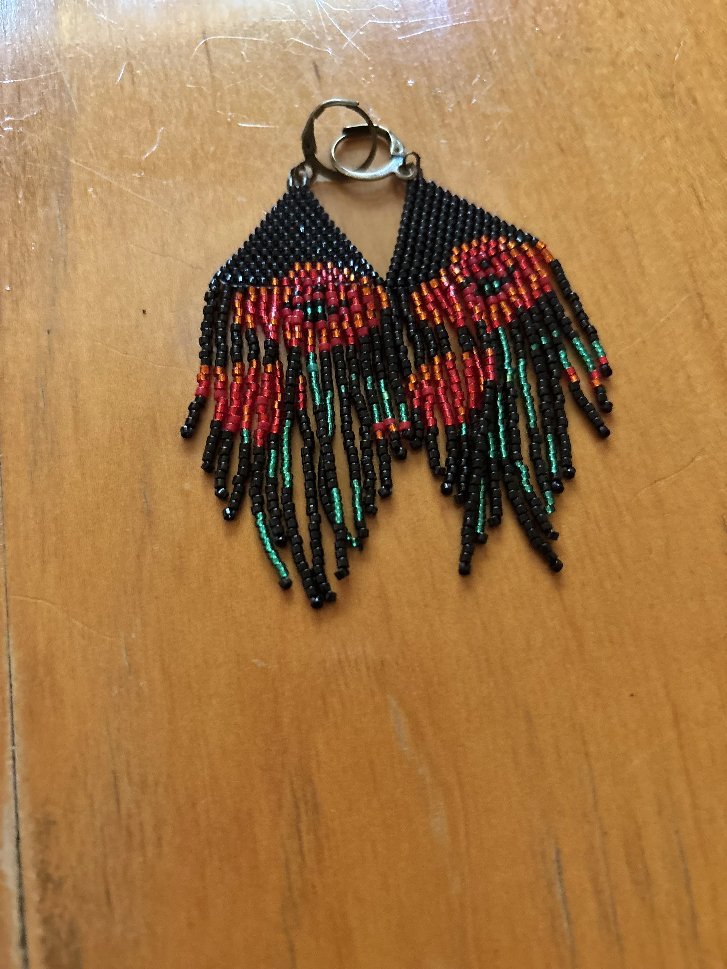 Beaded earrings with red, orange, black, and green beads, featuring a fringed design hanging from a silver clasp, placed on a wooden surface.