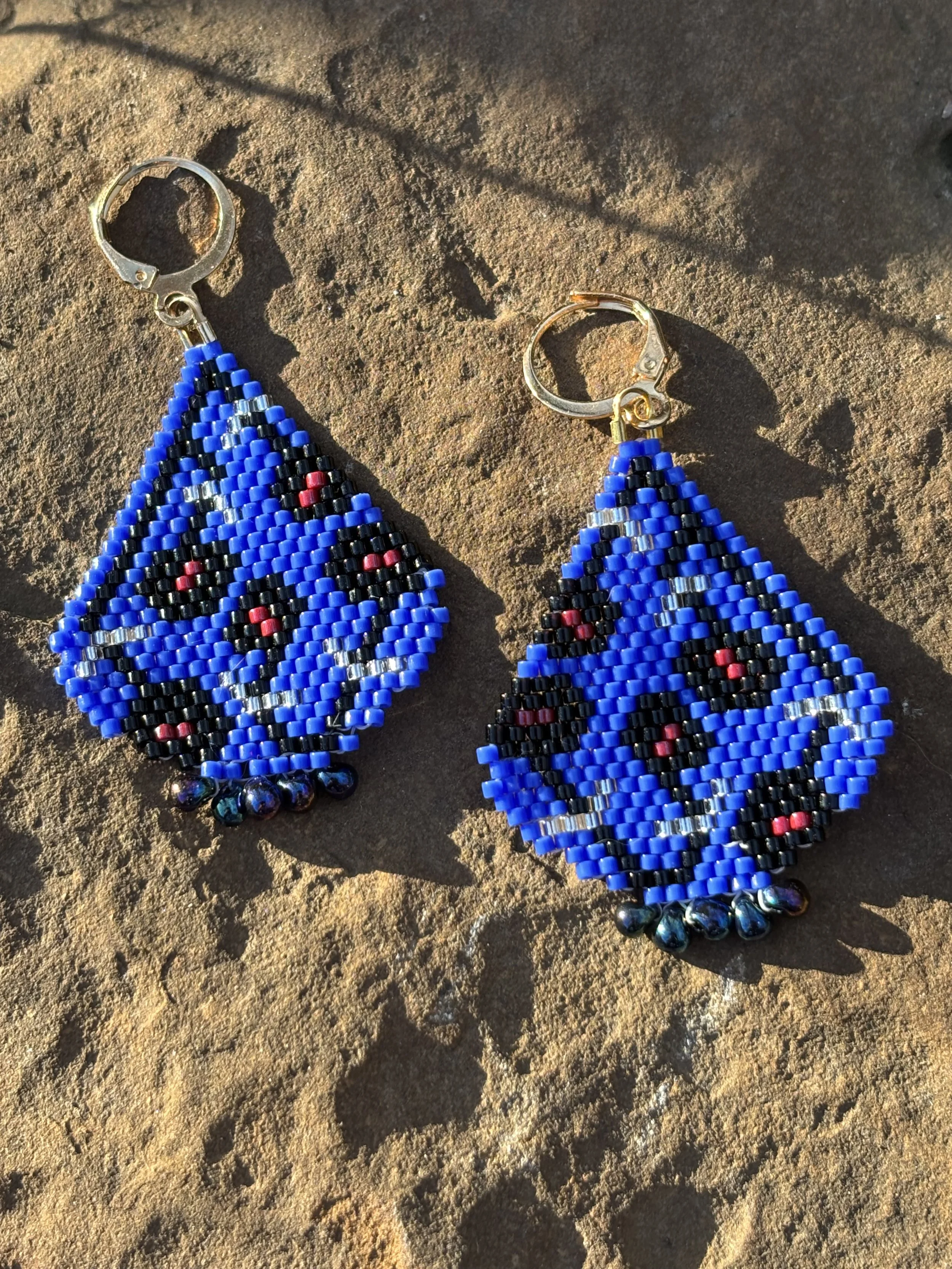 Beaded earrings with blue, black, silver, red, and metallic beads on a textured sandy surface with shadows.