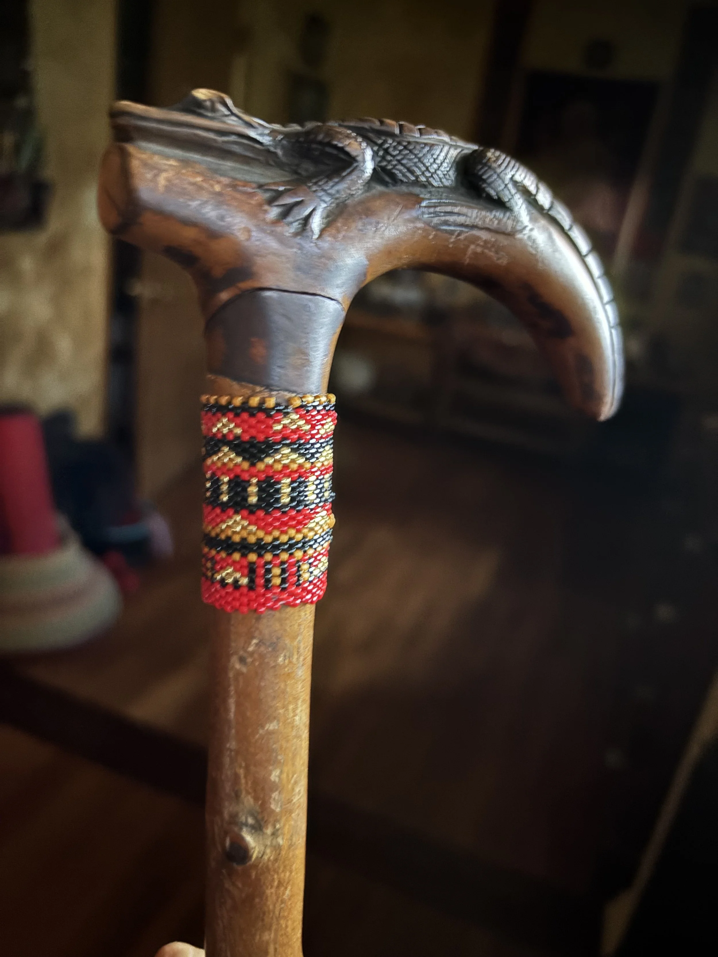 A carved wooden walking stick with a crocodile or lizard figure on top, decorated with a colorful beaded band around the handle. The background is dark and blurry.