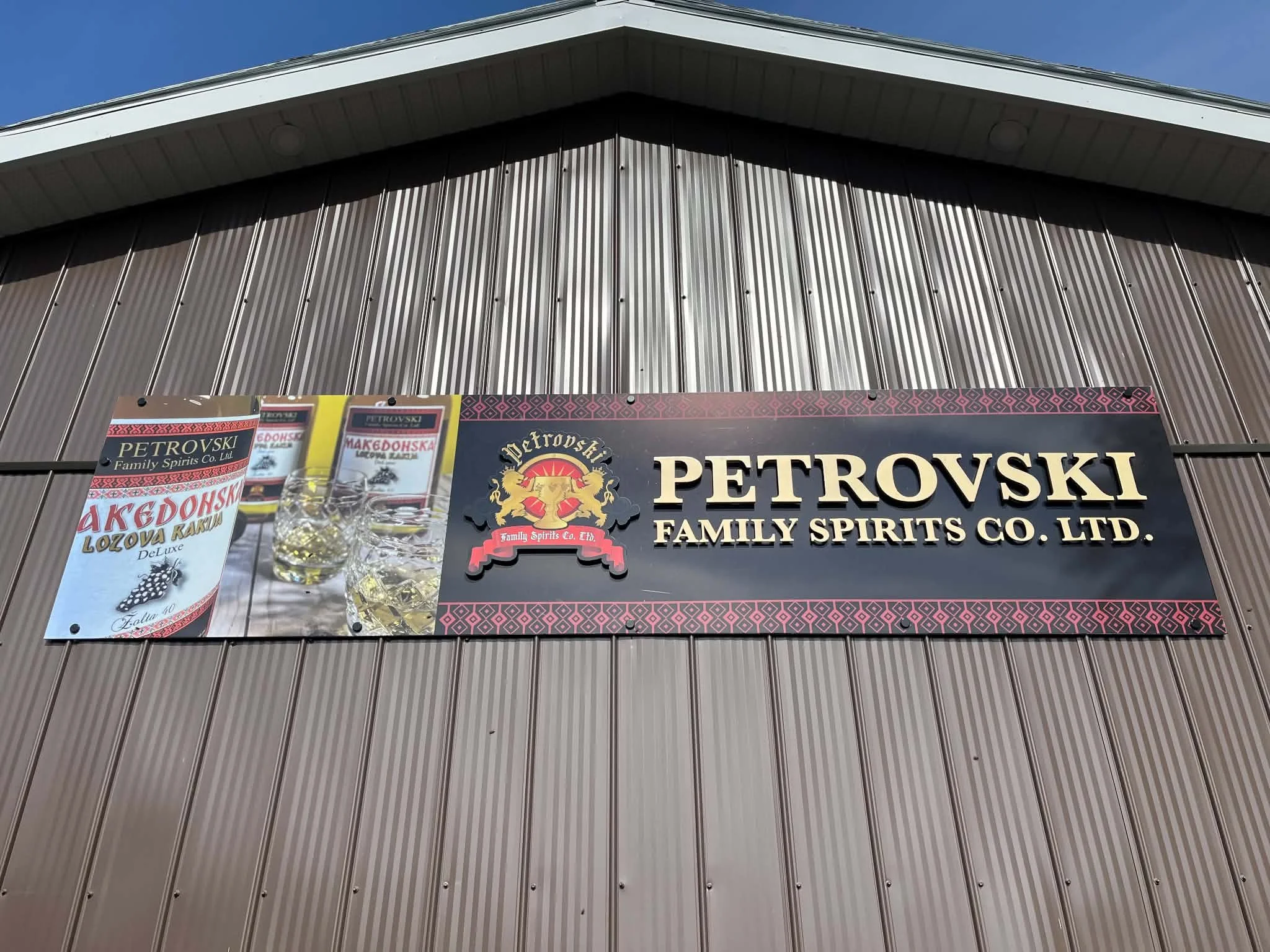 Exterior view of a building with a sign reading 'Petrovski Family Spirits Co. Ltd.' and promotional banners for Macedonian spirits. The building has vertical metal siding and a sloped roof.
