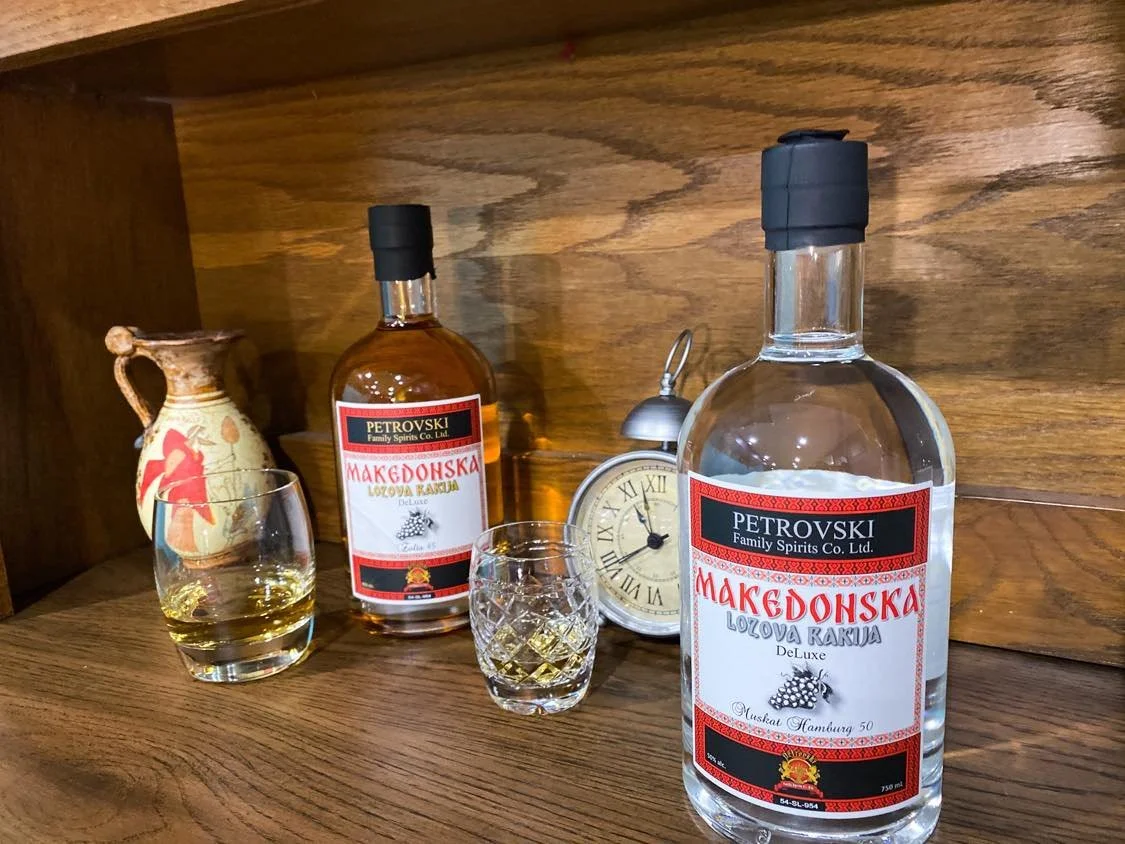 Two bottles of Macedonska brand spirits, two glasses filled with a golden liquid, a vintage clock, and a decorative vase on a wooden surface against a wood-paneled background.