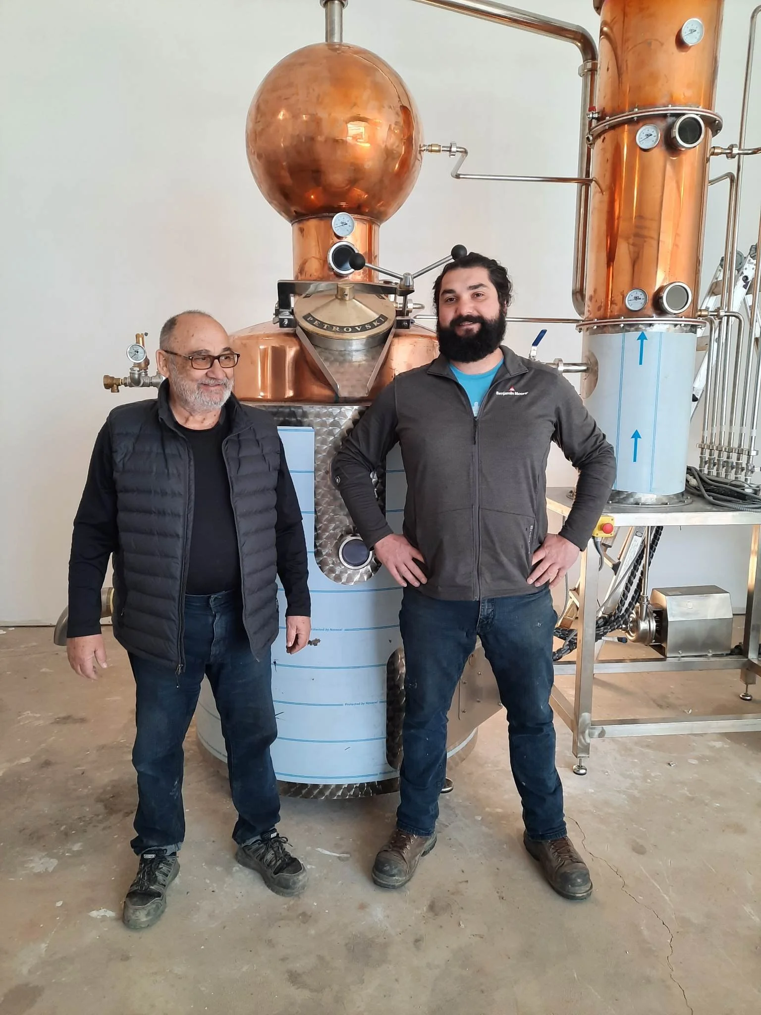 Two men standing in front of a large copper still used for distillation, with one man smiling and wearing glasses, and the other with a beard smiling and hands on hips.