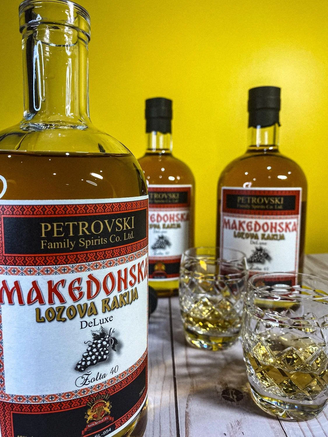Three bottles of Macedonian brandy on a wooden table with two glasses filled with the drink in front of a yellow wall.