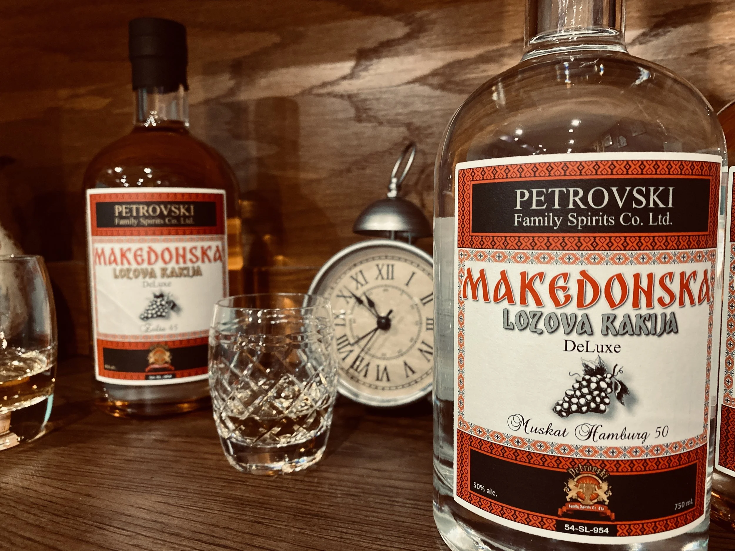 A bottle of Petrovsky Macedonian Lozova Rakija, a clear spirit, on a wooden table with a decorative clock and glasses nearby.