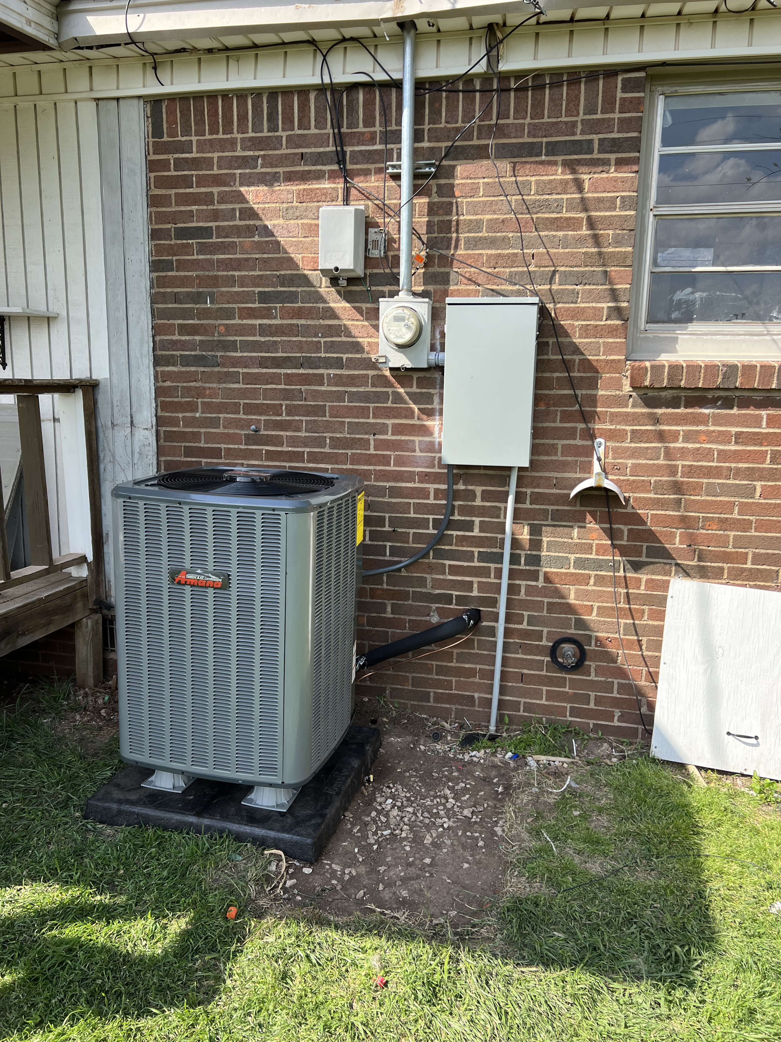 Outdoor view of a brick wall with an air conditioning unit, electrical meter, and various wires mounted on it, next to a window and a partially visible white door. Grass and dirt are in the foreground.
