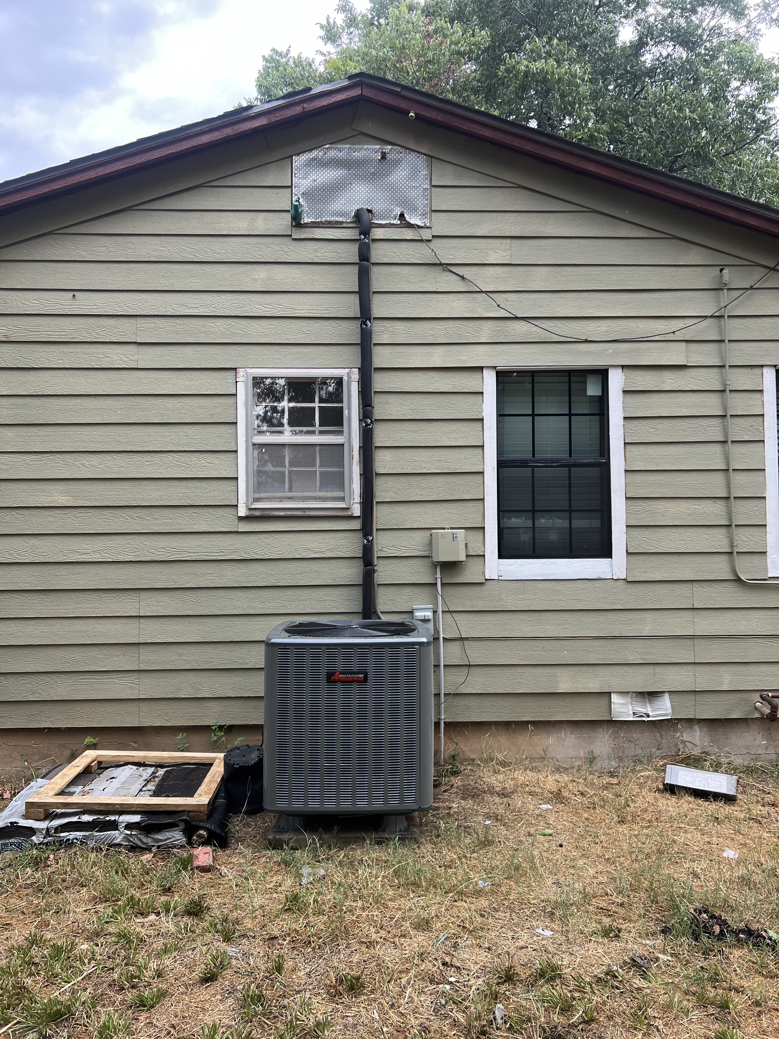 Backyard with beige house siding, two windows, and an air conditioning unit. Items on the ground include a wooden frame, black pipes, and miscellaneous debris. Tree branches are visible above the house.