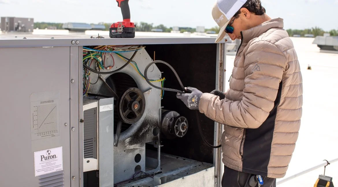 A technician working on HVAC equipment on a building rooftop during daytime. The technician is wearing a beige jacket, gloves, sunglasses, and a cap, and is using a tool to perform maintenance or repairs.