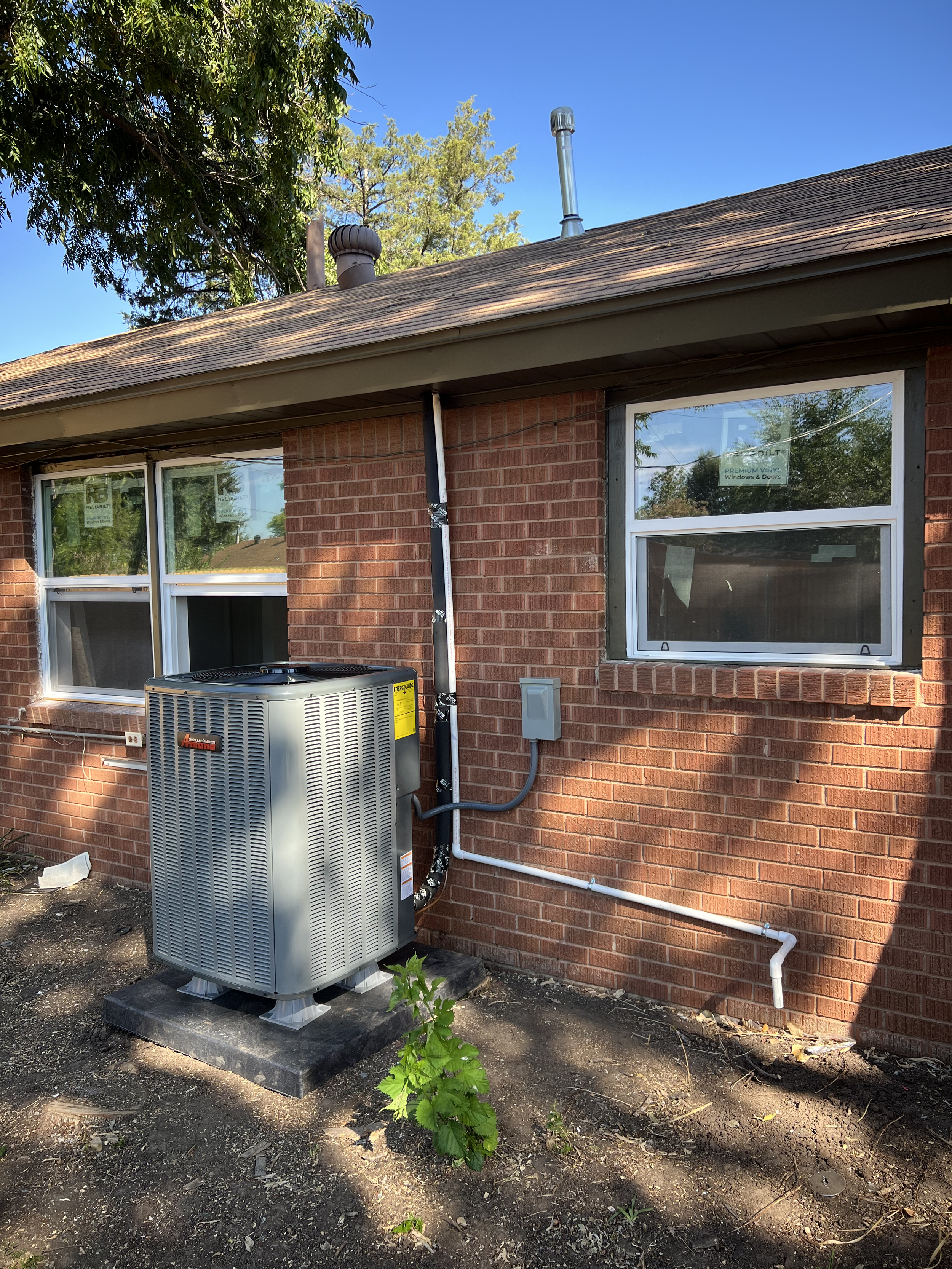 The image shows the side of a brick house with two windows. There is an HVAC unit installed on a concrete pad on the ground, with some pipes and wiring attached to the house. The area around the HVAC unit has dirt and small plants, and there are tree