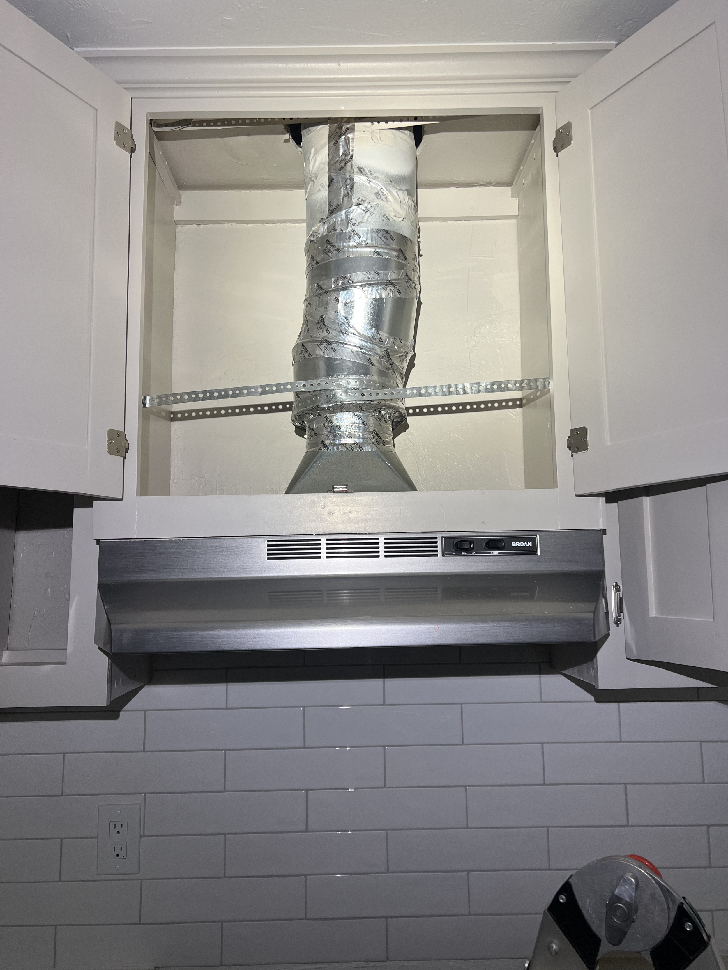Kitchen range with exhaust hood, cabinets open revealing ductwork with metal foil insulation and ridged metal duct, white tiled wall, electrical outlet underneath, and a step ladder in front.