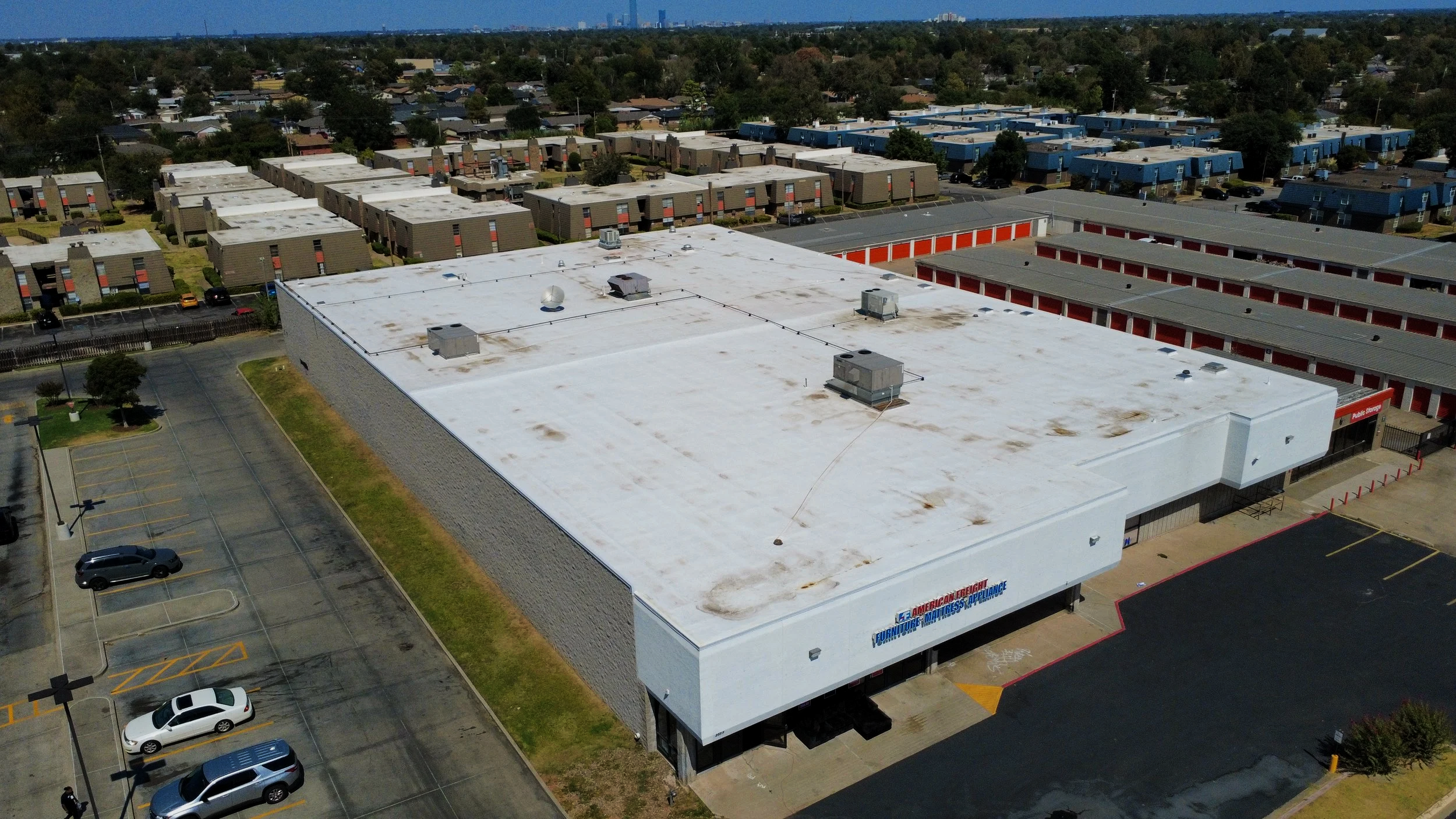 An aerial view of a large retail store with a white flat roof, parking lot with a few cars, and surrounding residential neighborhood with houses and trees.