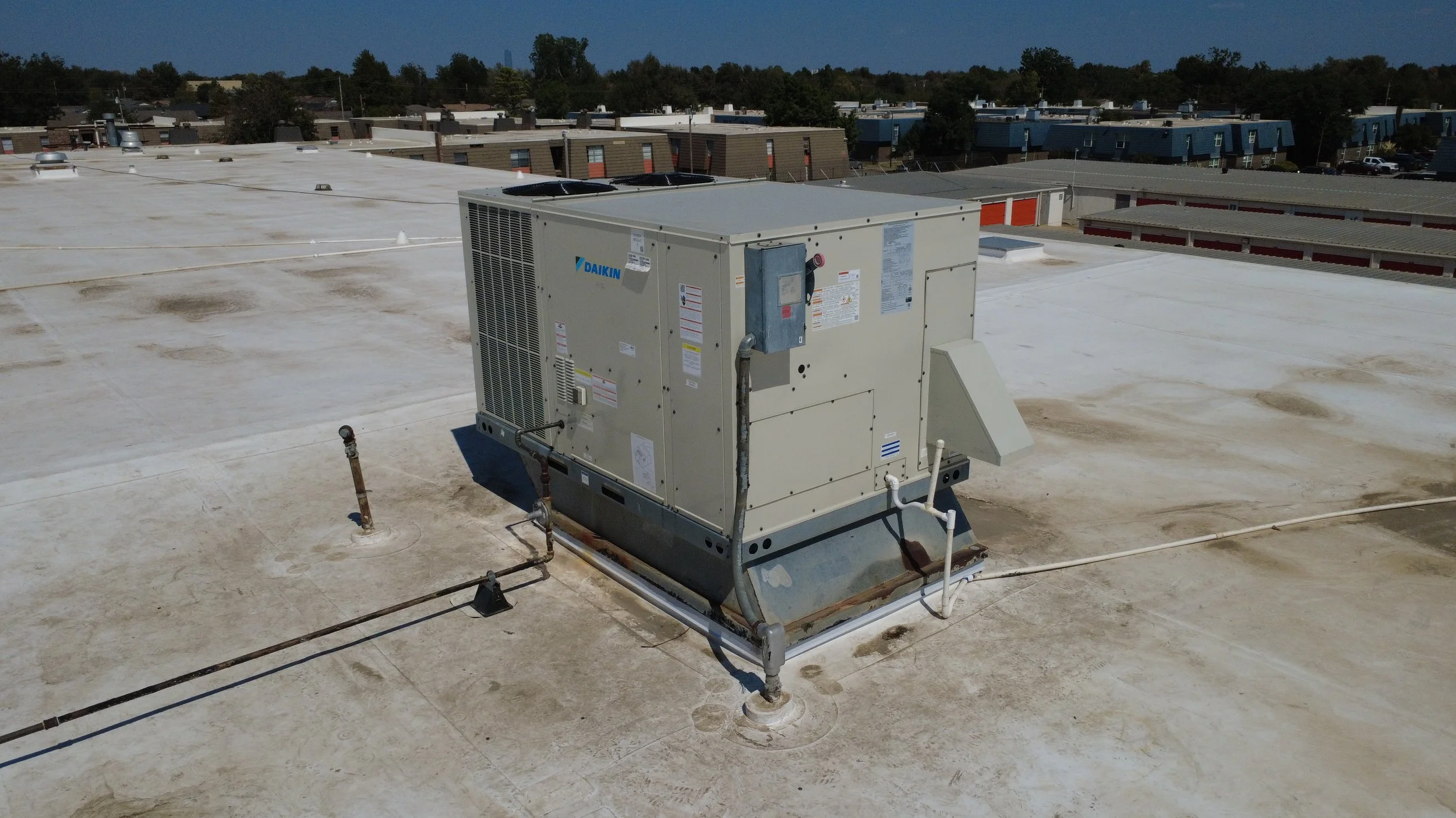 Rooftop with a large HVAC unit and surrounding pipes, residential neighborhood in the background under a clear sky.