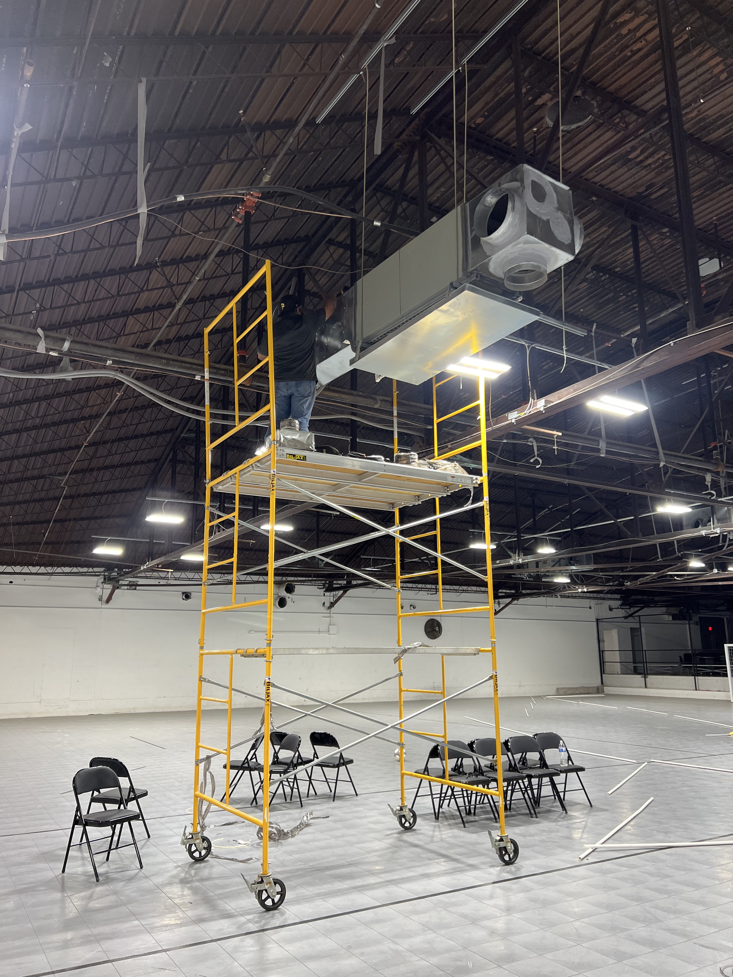 A worker on a yellow scaffolding installs or maintains a large metal HVAC air handling unit ceiling duct in an indoor space that appears to be an industrial or commercial building. The room has a high ceiling with exposed metal beams and black piping