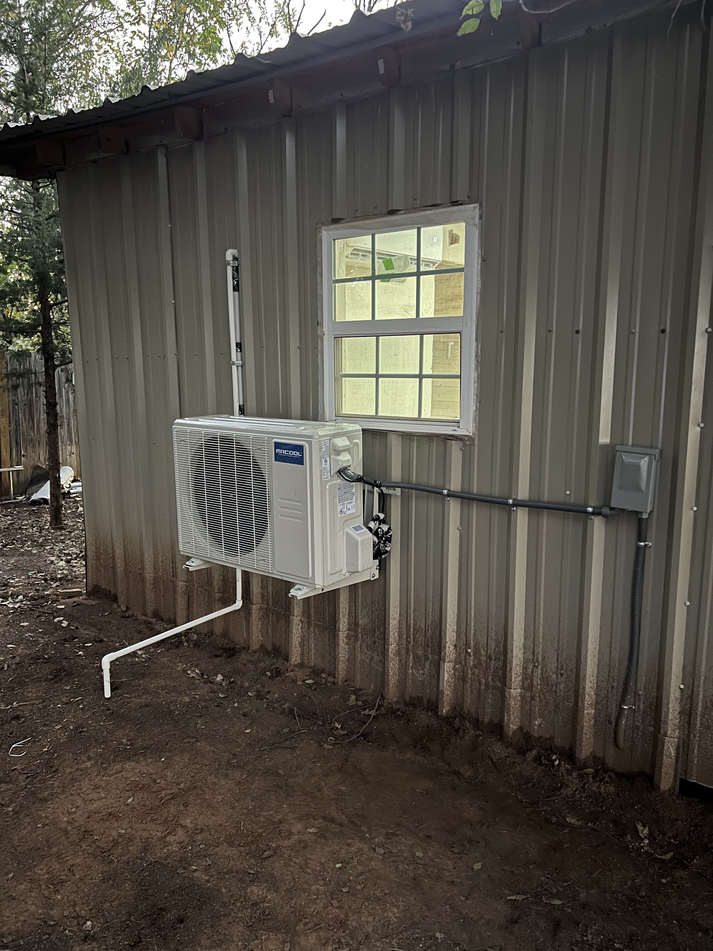 The exterior of a metal shed or building with a small window, an air conditioning unit, and pipework attached, with dirt ground in front.