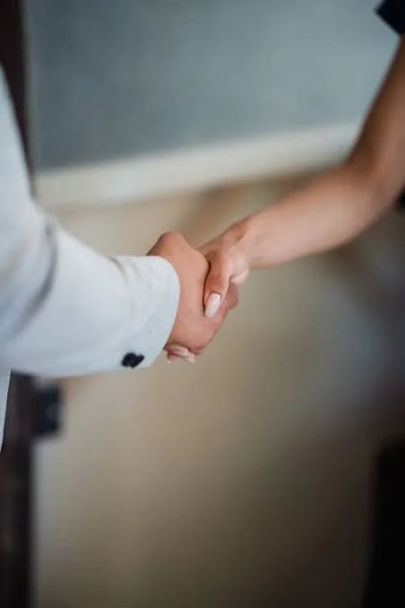 Close-up of two people shaking hands, one in a white blazer and the other with a short-sleeved shirt, against a neutral background.