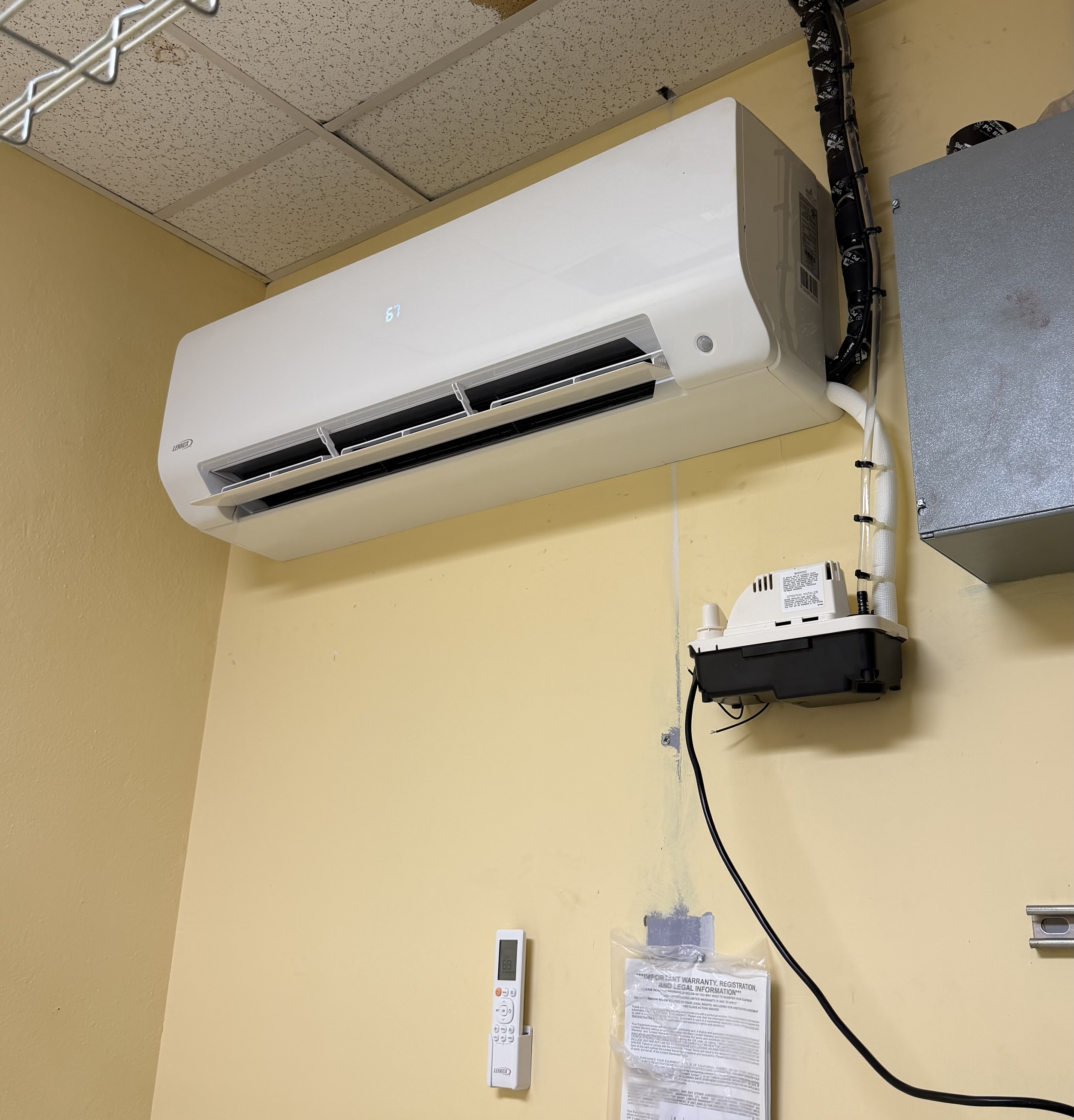 An air conditioning unit mounted on a yellow wall with digital display reading 67, a white remote control mounted below, a paper, and a black box with cables connected to it.