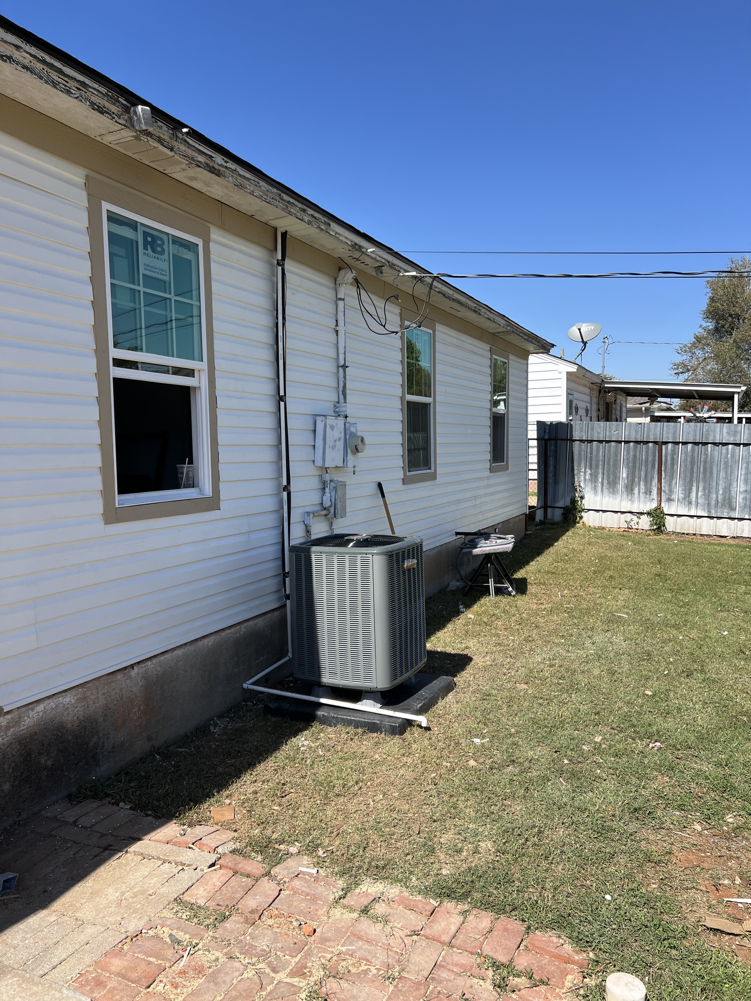 Backyard view showing the side of a house with white siding, a window, and an air conditioning unit on the ground. There is a brick pathway and a small patch of grass, with a wooden fence and some trees in the background under a clear blue sky.