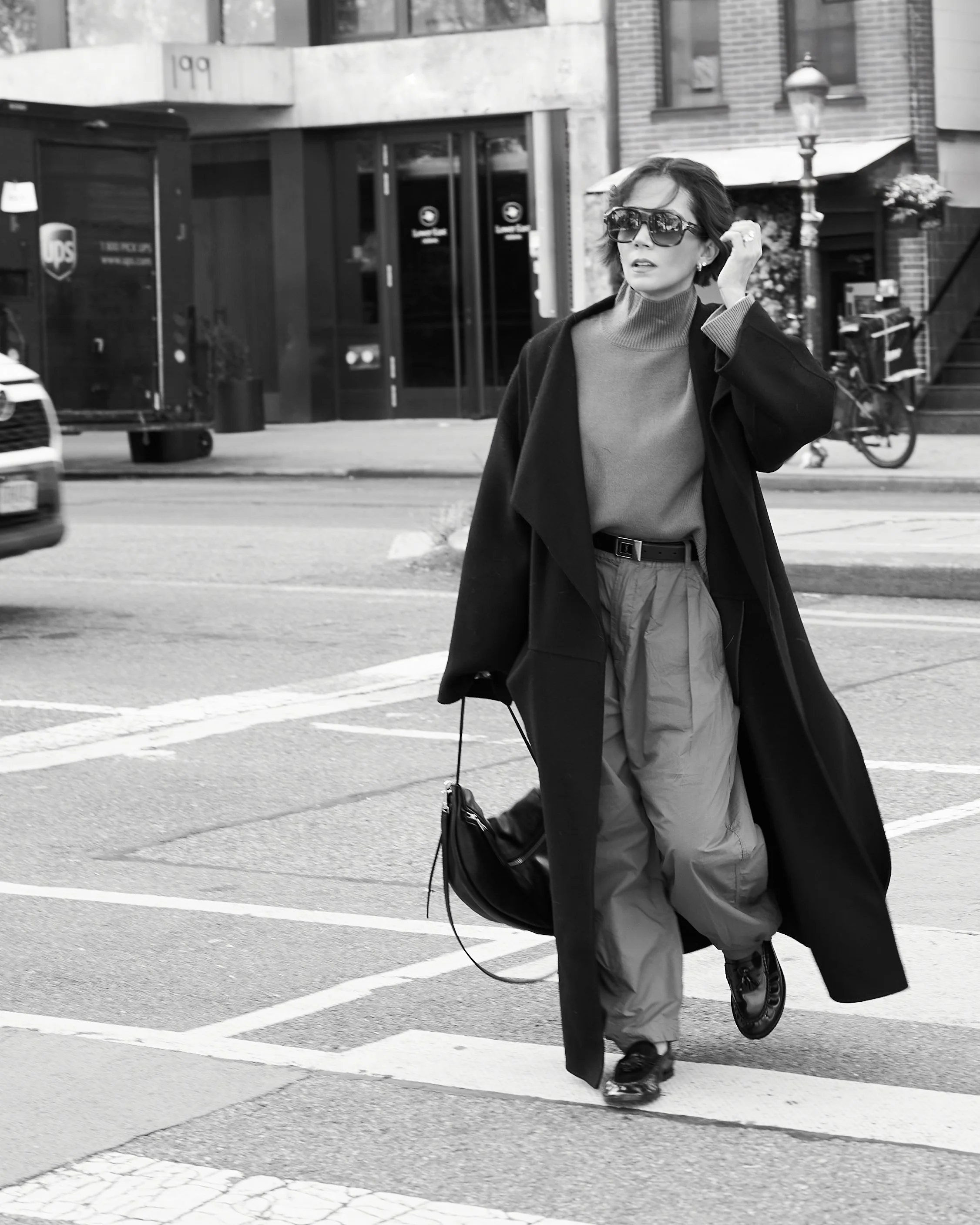 Black and white street style photo of a long black coat, trousers, sunglasses and a shoulder bag