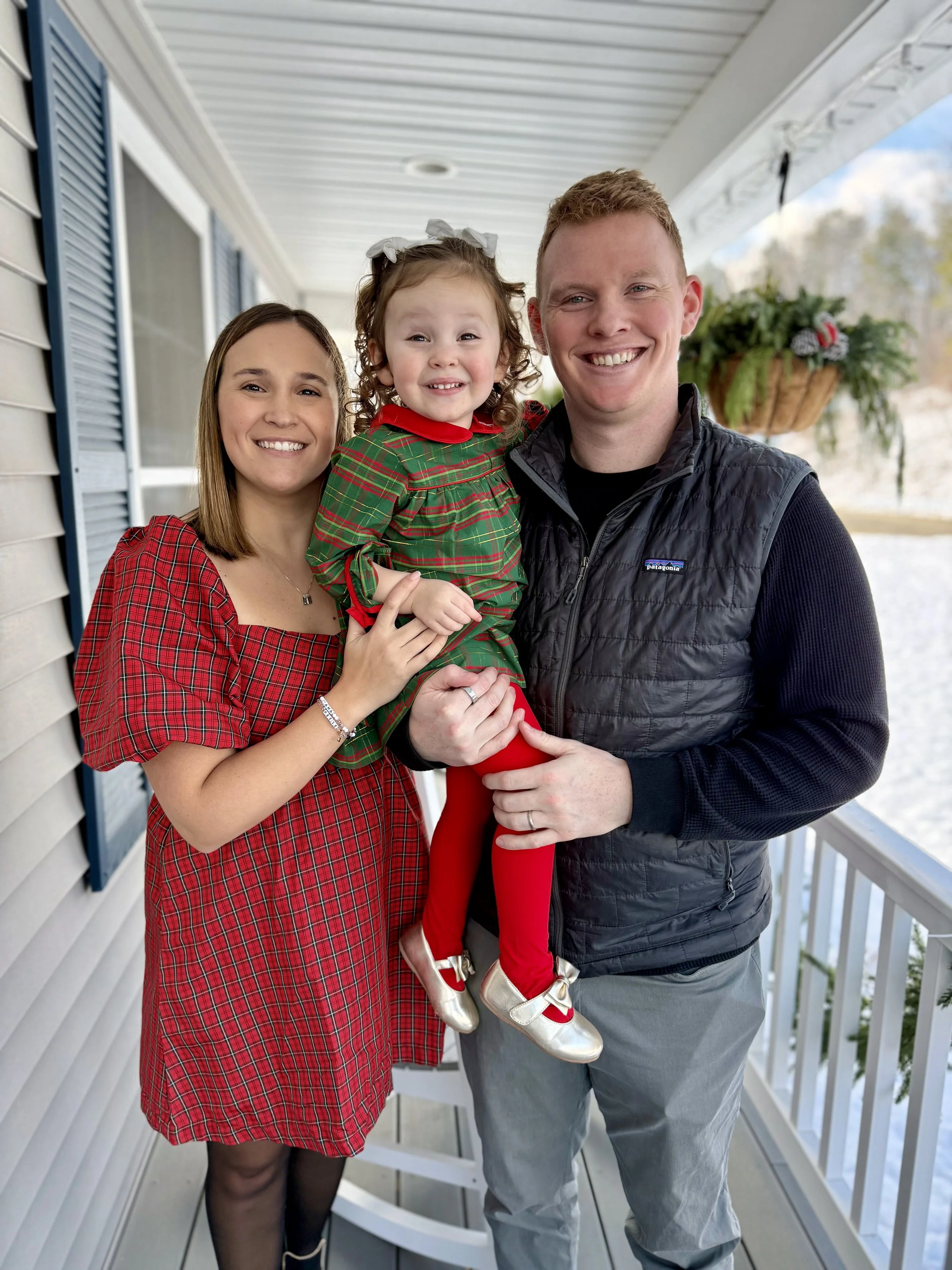 Family of three on porch, woman in red plaid dress, young girl in green plaid dress and ballet shoes, man in black jacket.