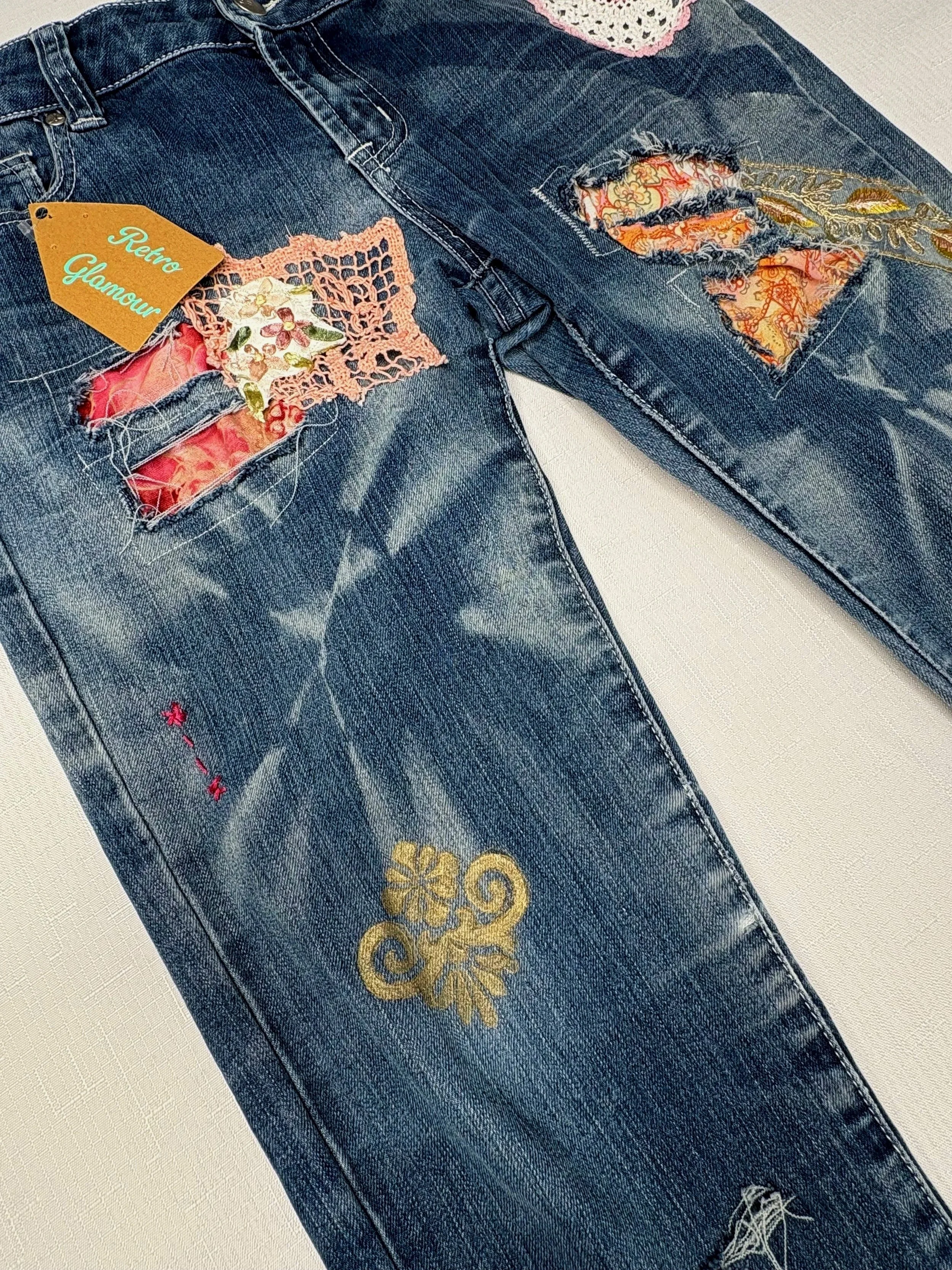 Close-up of dark denim overalls with floral fabric patches, pink crocheted trim, embroidery, and a retro glamour tag.