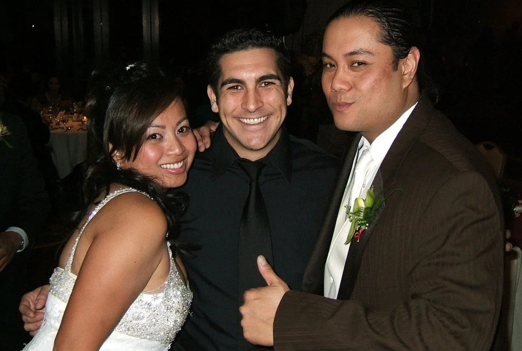 Three people smiling and posing together at a formal event. The woman on the left is wearing a white dress with spaghetti straps. The man in the middle is dressed in a black shirt and tie. The man on the right is in a brown suit with a boutonniere and white shirt.