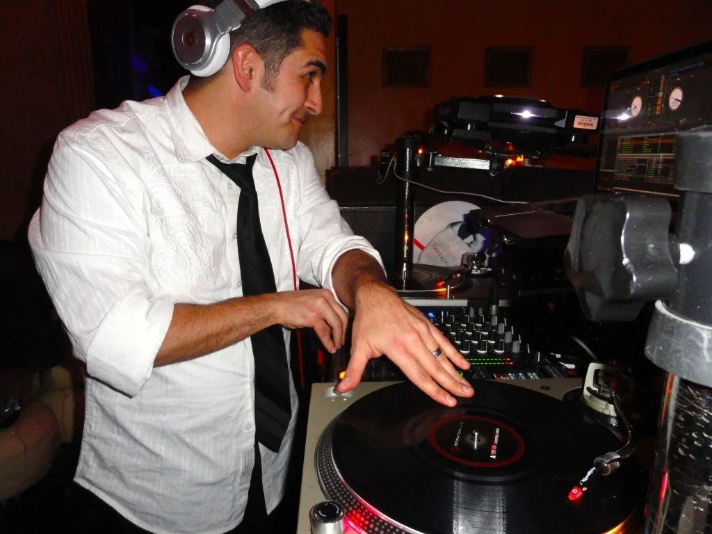 A young man wearing headphones and a white dress shirt with rolled-up sleeves and a black tie, smiling while DJing with a turntable and mixer at a party or event.