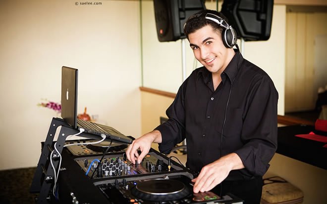 A young man wearing a black shirt and headphones mixes music on a DJ controller with a laptop in front of him, smiling at the camera.