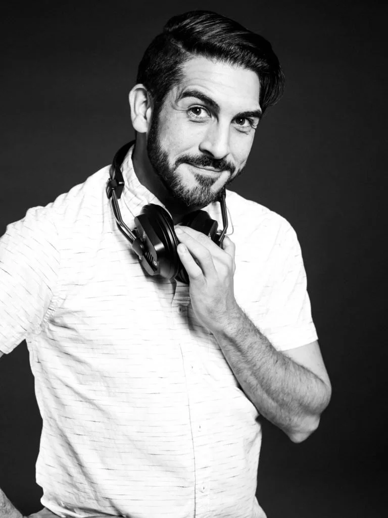 A young man with styled dark hair and a beard, smiling confidently, wearing a light-colored striped shirt and headphones around his neck, posed against a dark background.