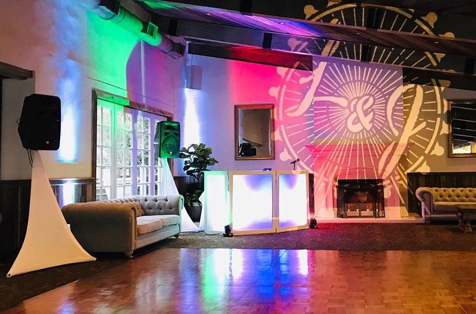 Indoor event space with colorful lighting, a DJ booth, vintage sofas, a fireplace, and a large clock projection on the wall.