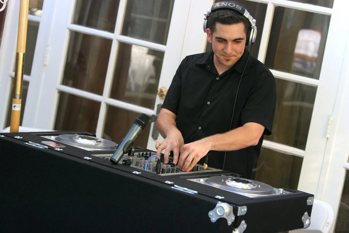A man with dark hair wearing headphones and a black shirt is DJing at a table with turntables and a microphone in front of glass doors.