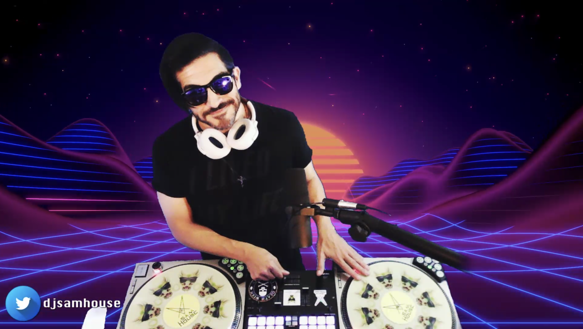 A DJ with dark hair, sunglasses, and a beard wearing a black t-shirt and headphones around his neck, is standing behind a DJ controller with neon-lit turntables. The background features a digital, retro-futuristic landscape with neon purple and pink rolling hills, a setting sun, and a starry sky, creating a 1980s cyberpunk aesthetic.