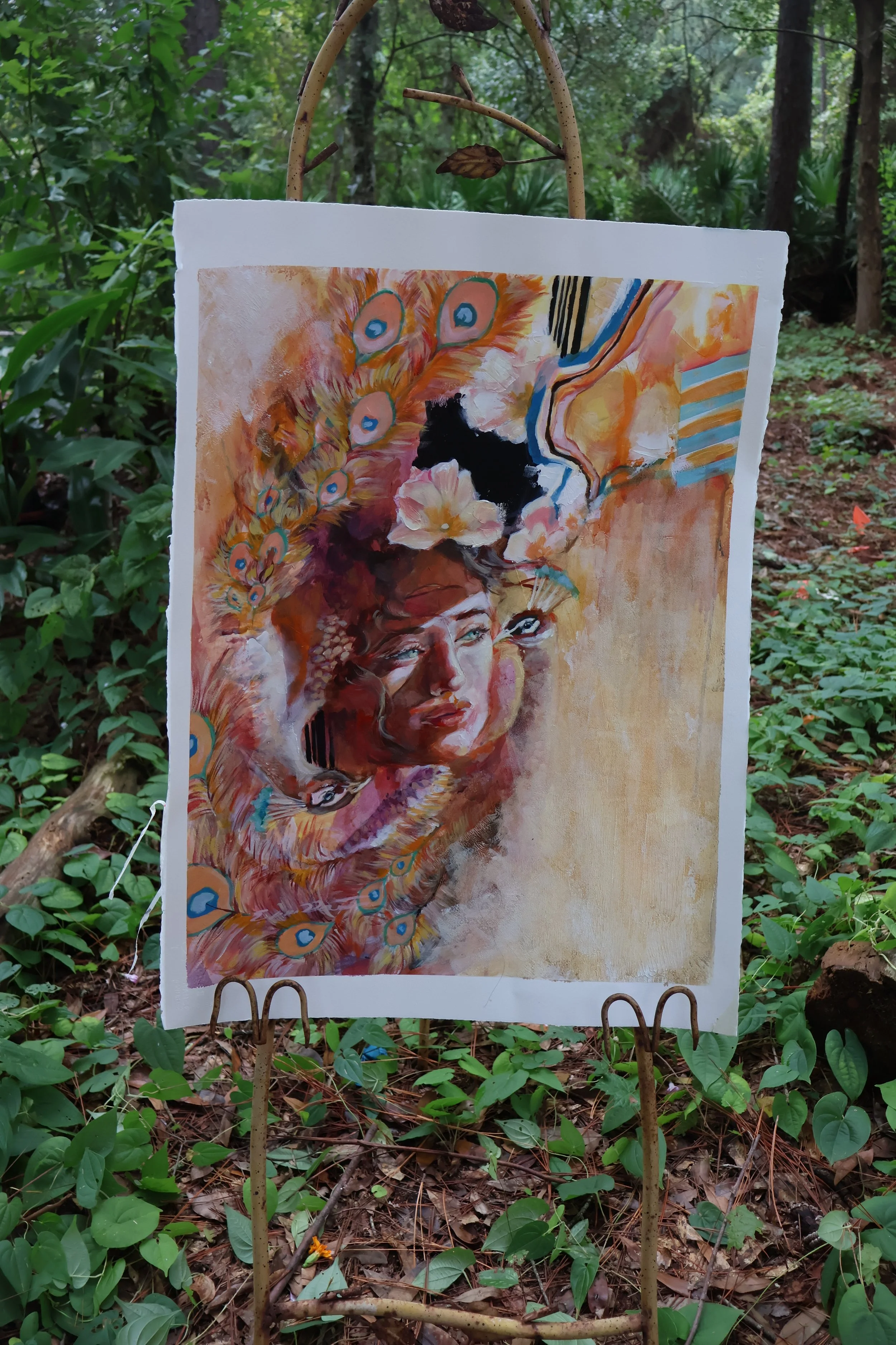 Colorful abstract painting featuring a woman's portrait, peacock feathers, and flowers displayed on an easel in a wooded setting.