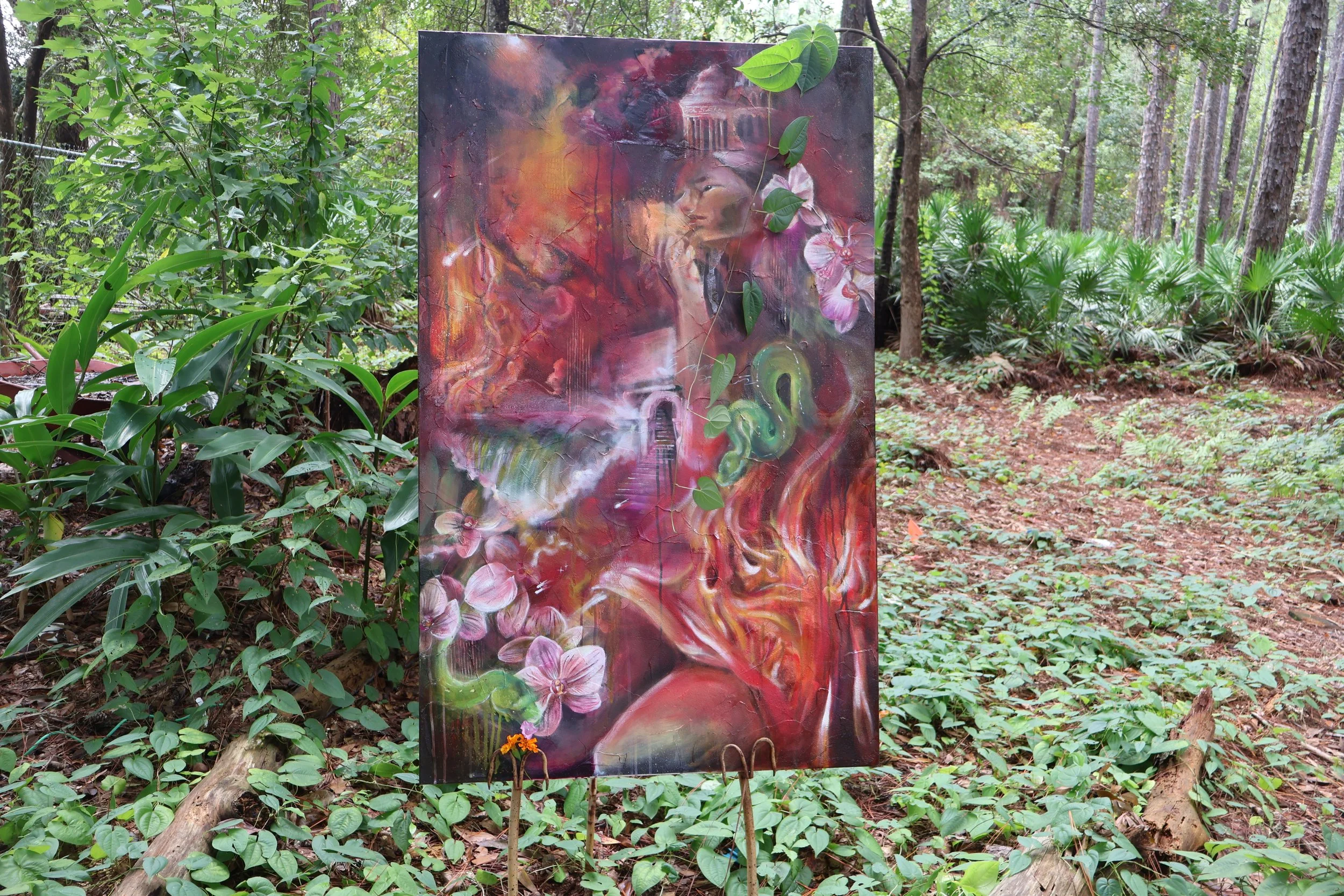 Colorful mixed-media painting displayed on an easel in a lush forest, featuring a woman's portrait, flowers, a snake, and architectural imagery.