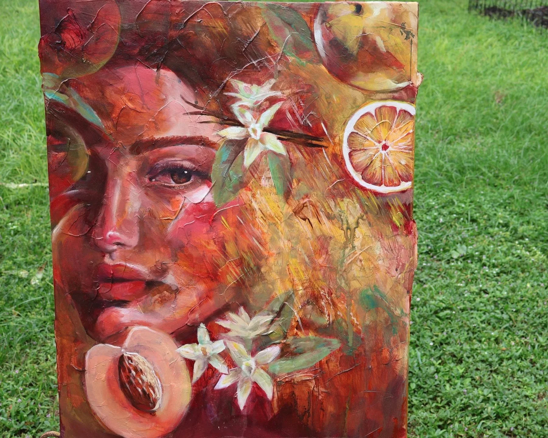 Colorful abstract painting of a woman surrounded by white flowers, citrus slices, and a halved pomegranate, displayed outdoors on grass
